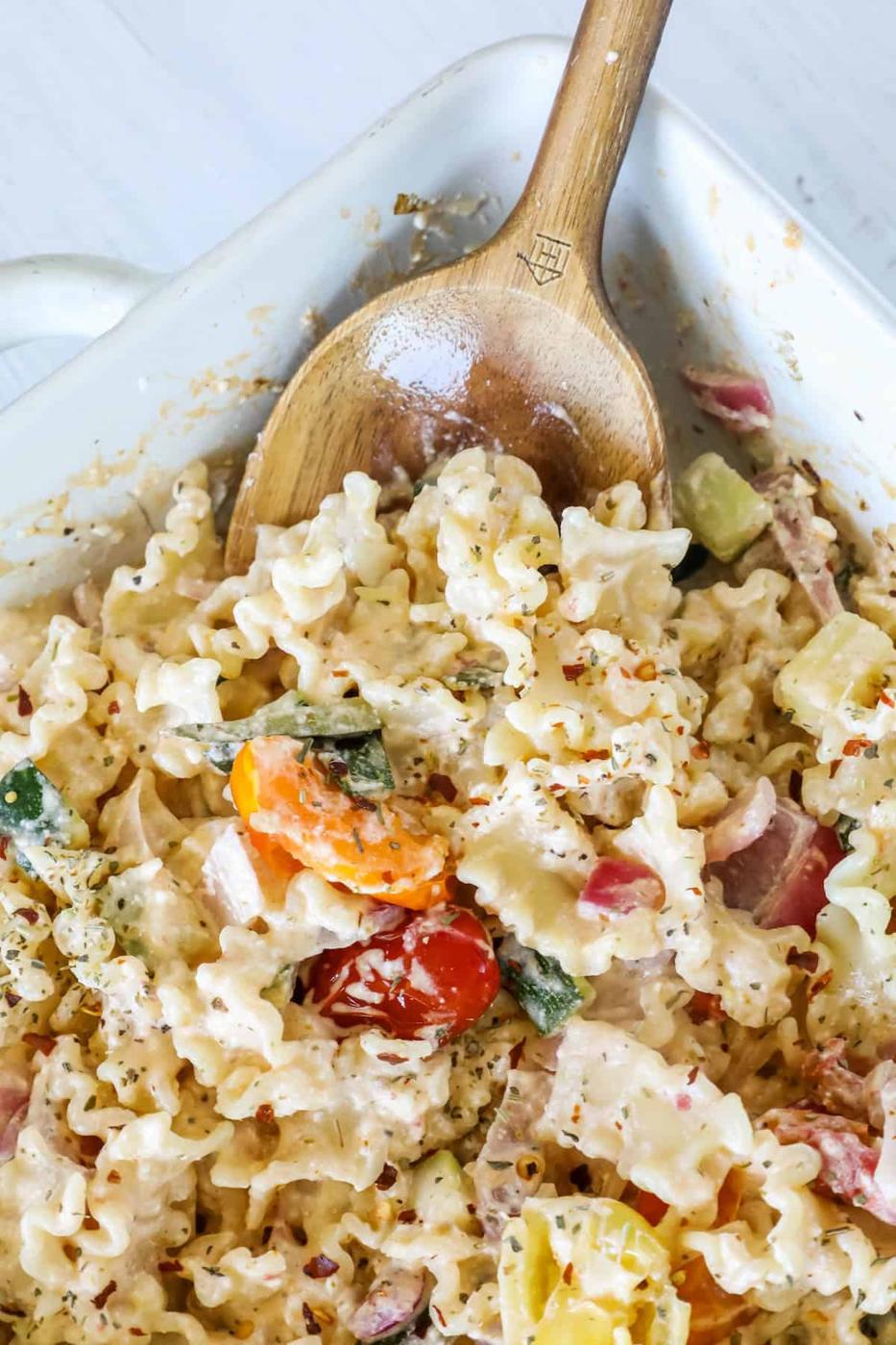Close-up of a pasta dish with a creamy sauce, mixed with vegetables such as tomatoes, zucchini, and bell peppers, served in a white casserole dish with a wooden spoon.