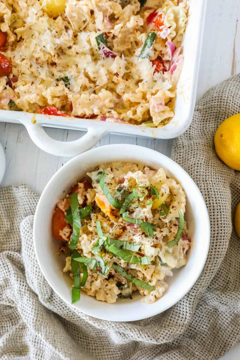 A bowl of pasta topped with chopped basil sits on a textured cloth next to a baking dish with more of the same pasta. Two lemons rest nearby on a white wooden surface.