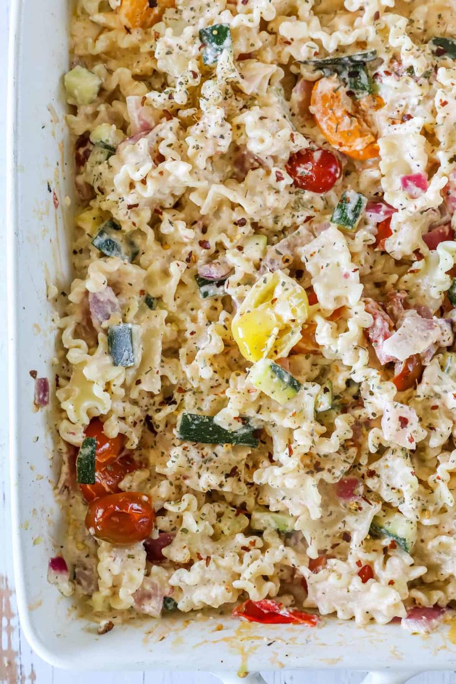 A close-up view of a pasta salad with mafaldine noodles, cherry tomatoes, zucchini, red onion, and a creamy dressing in a white dish.