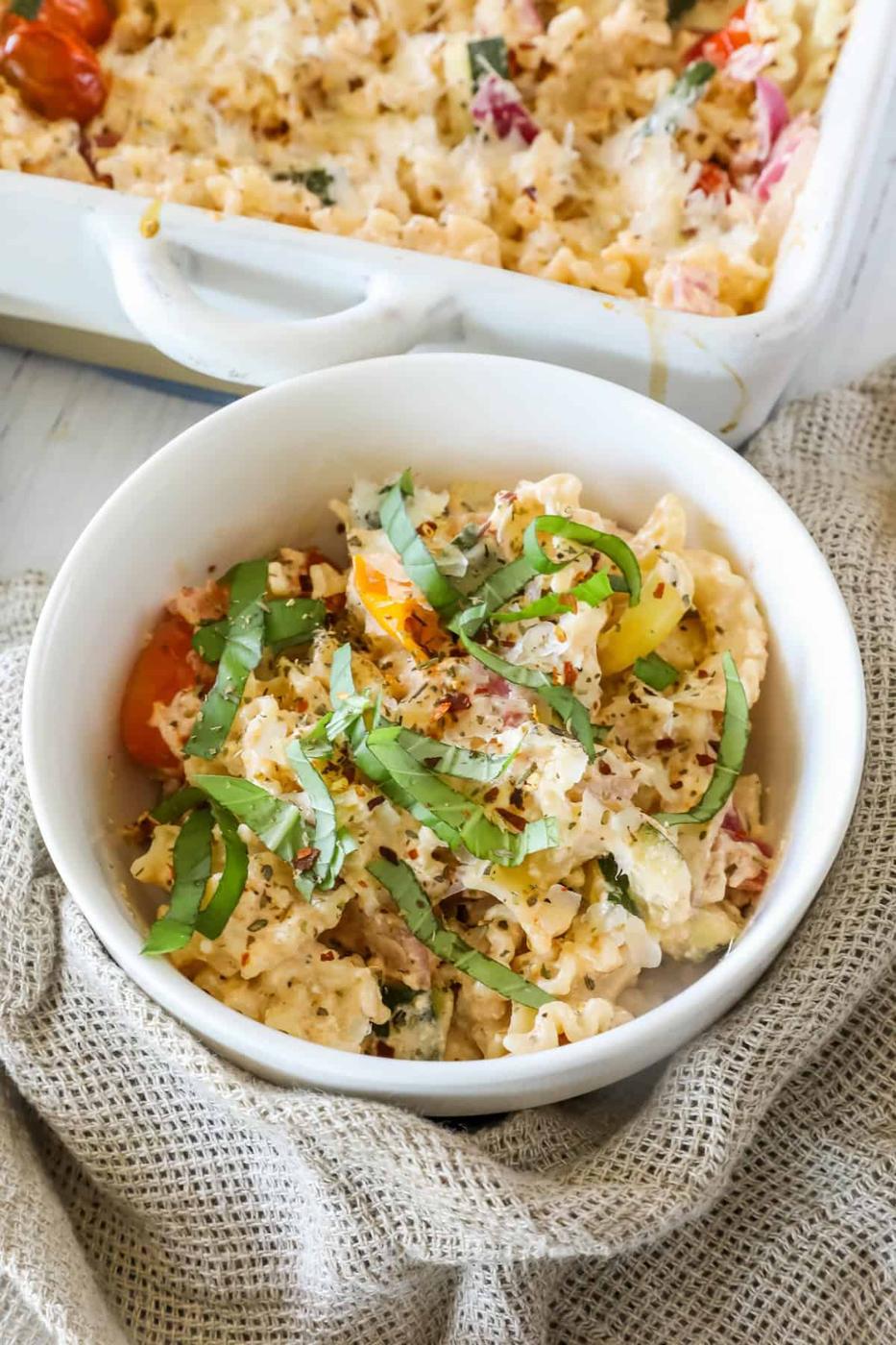A white bowl of baked pasta mixed with vegetables, topped with fresh basil, sits on a textured cloth near a baking dish containing more pasta.