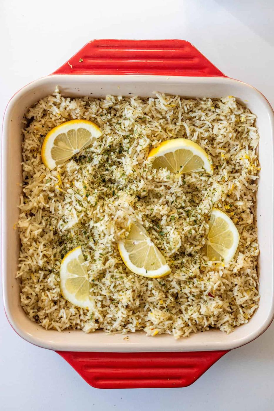A square red baking dish filled with Baked Lemon Rice, garnished with fresh lemon slices and green herbs.