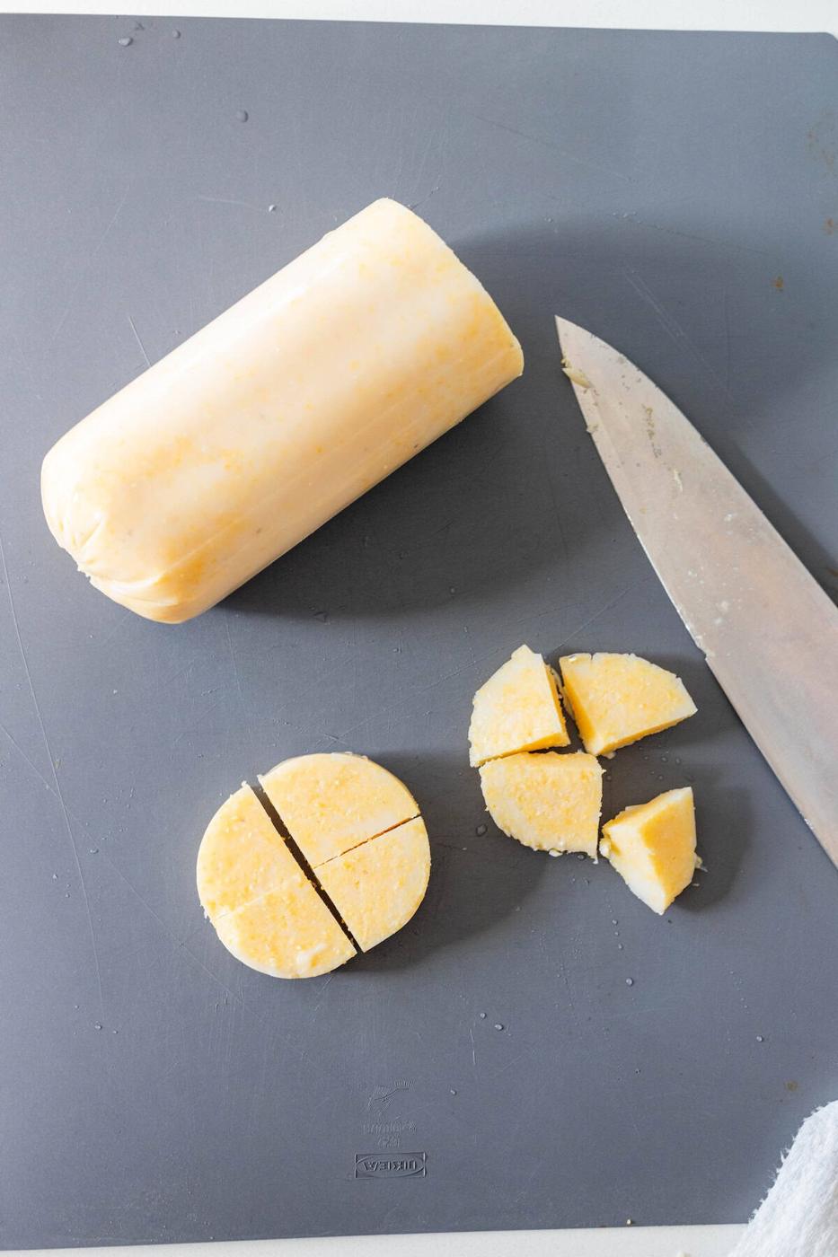 A log of dough being sliced with a knife on a dark cutting board; some circular pieces, perfect for baked appetizers or polenta bites, are also cut into quarters.