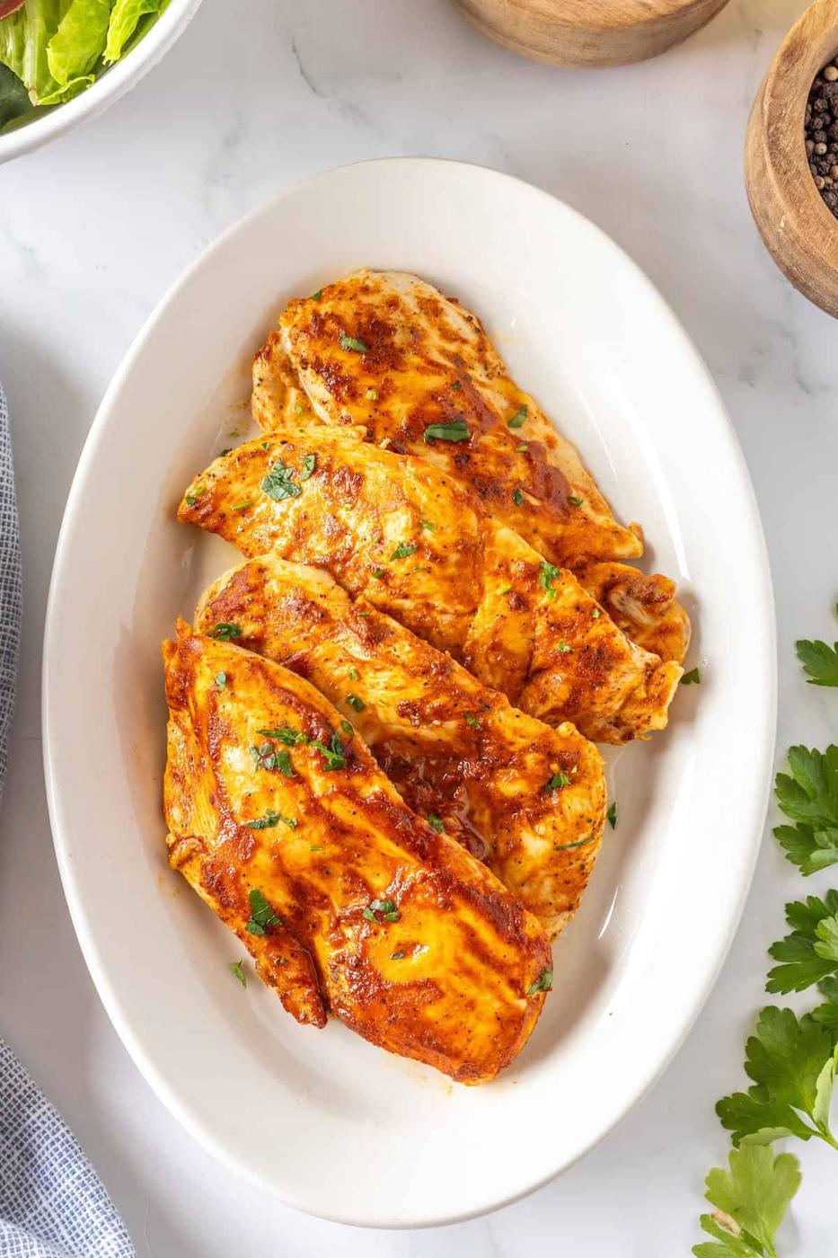 Thin baked chicken breasts seasoned with herbs and spices rest elegantly on a white oval plate, garnished beautifully with fresh parsley.