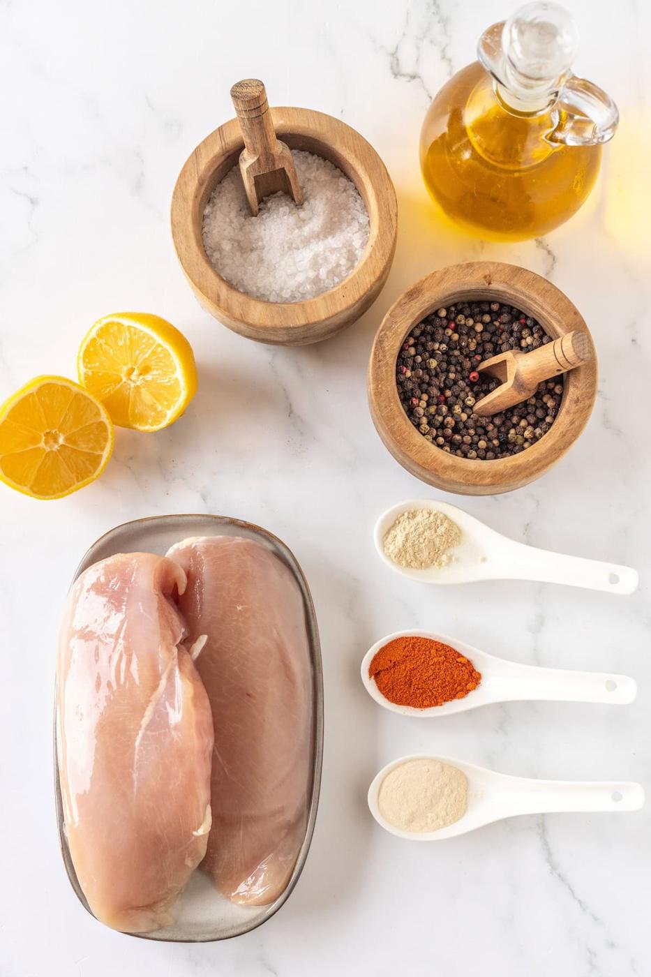Raw chicken breasts, lemon halves, and olive oil rest in a wooden bowl, accompanied by a sprinkle of salt and peppercorns. Three white spoons hold an array of spices on the marble surface, ready to transform these ingredients into a delectable thin baked chicken masterpiece.