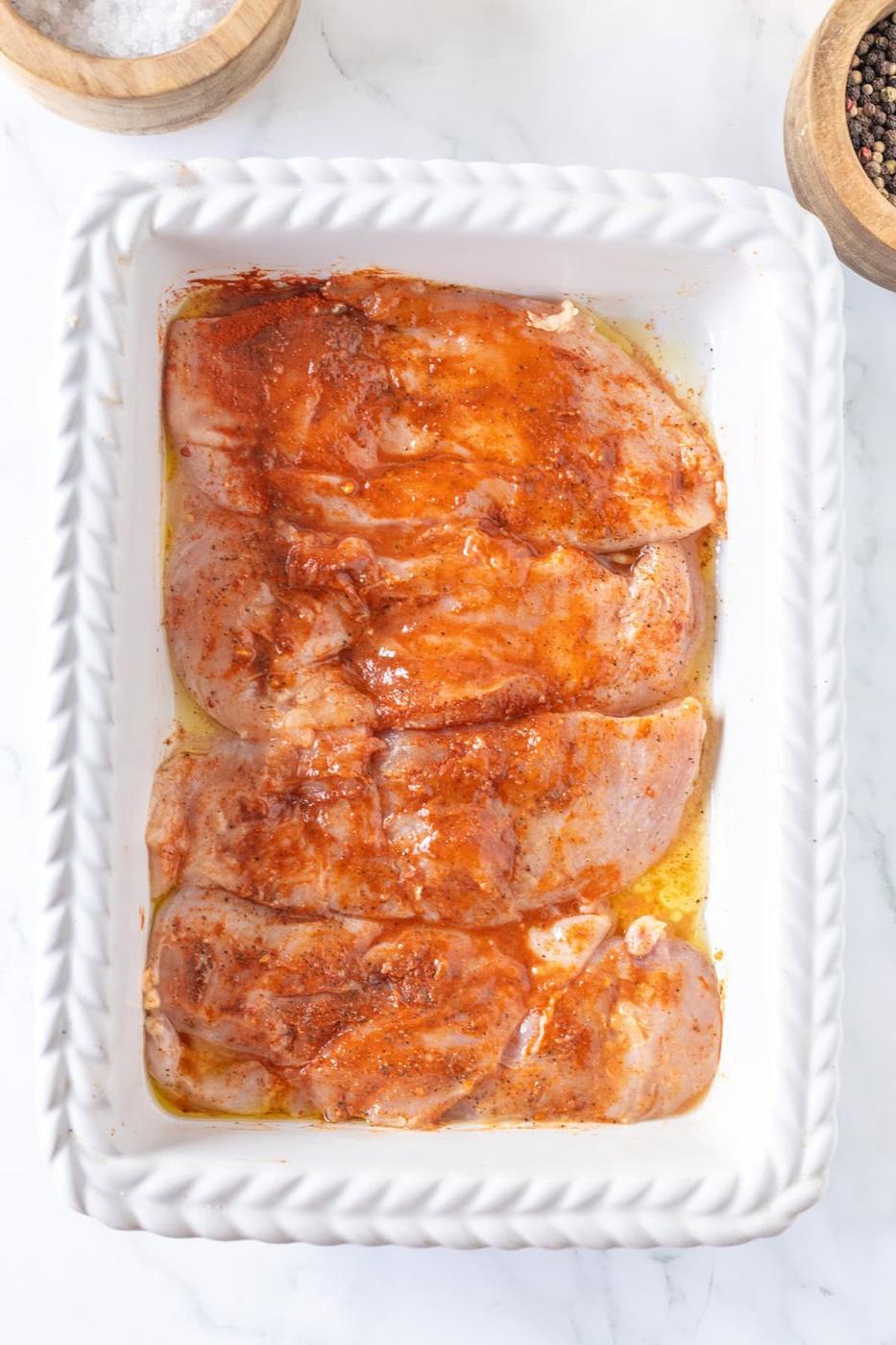 Thin chicken breasts are marinated with spices and oil in a white baking dish, ready to transform into one of your go-to chicken recipes, perfectly set on a marble surface.
