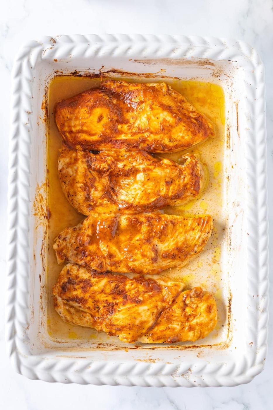 Four seasoned and thin baked chicken breasts rest perfectly in a white baking dish.