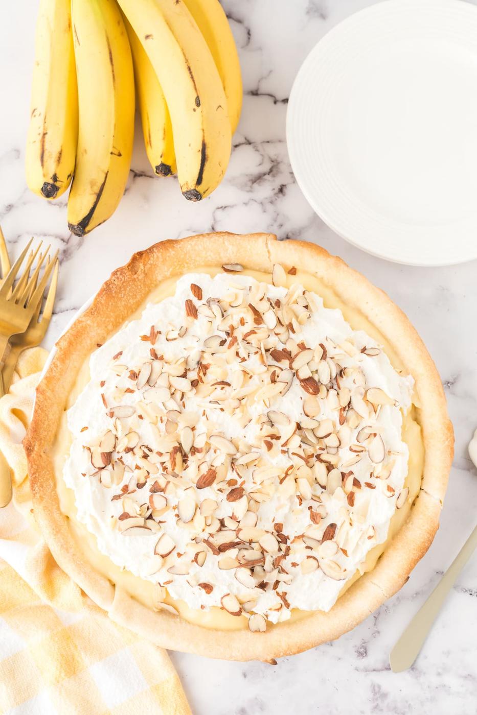 banana cream pie in a pie plate with almonds on top and bananas on a table