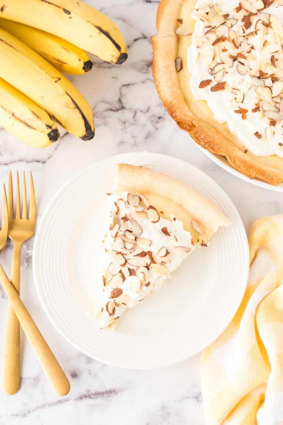 slice of banana cream pie on a plate with almonds on top and bananas on a table and a fork next to it