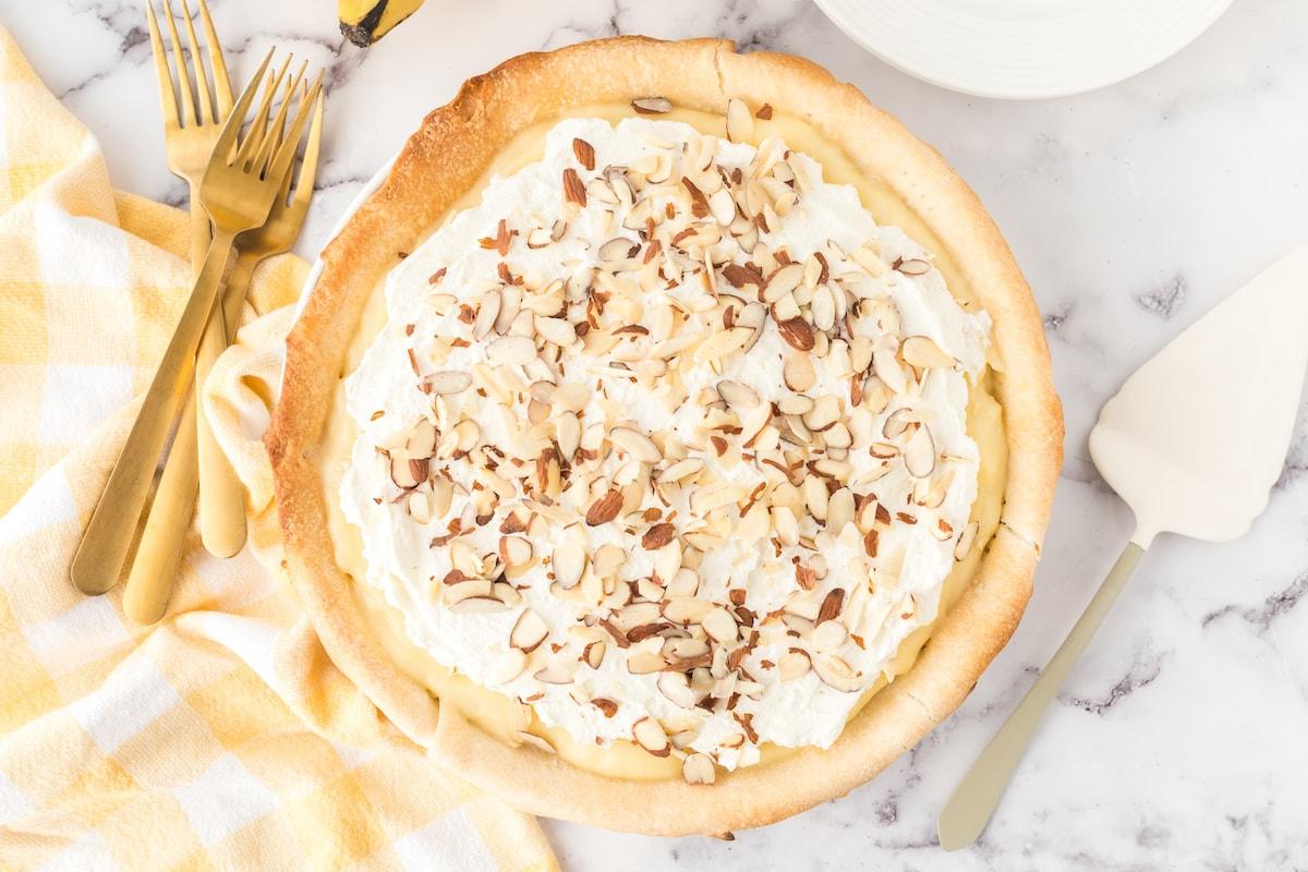slice of banana cream pie on a plate with almonds on top and bananas on a table with a pie knife next to it