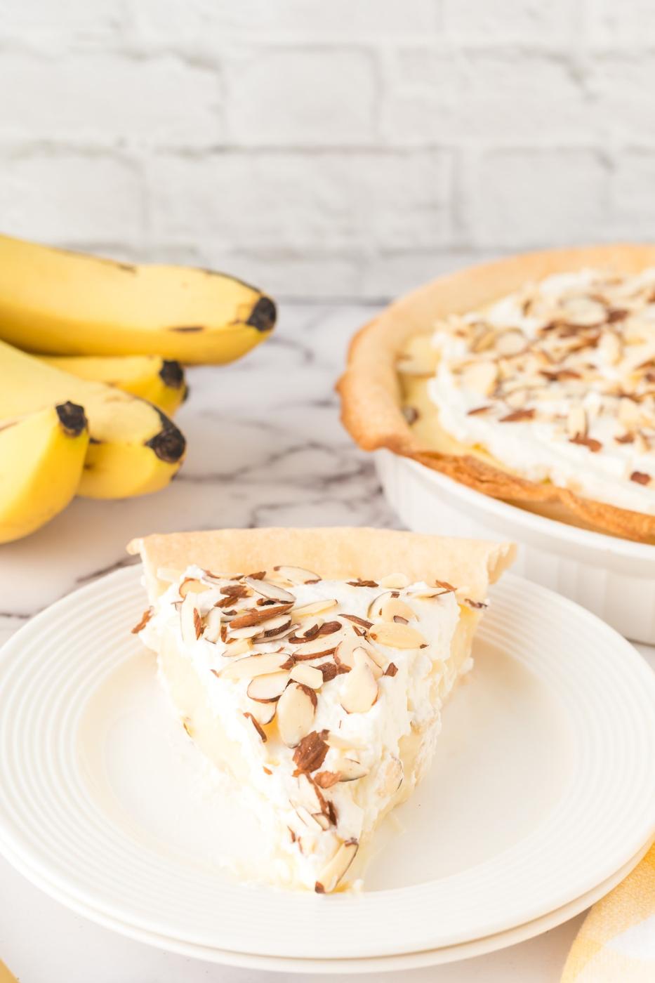slice of banana cream pie on a plate with almonds on top and bananas on a table