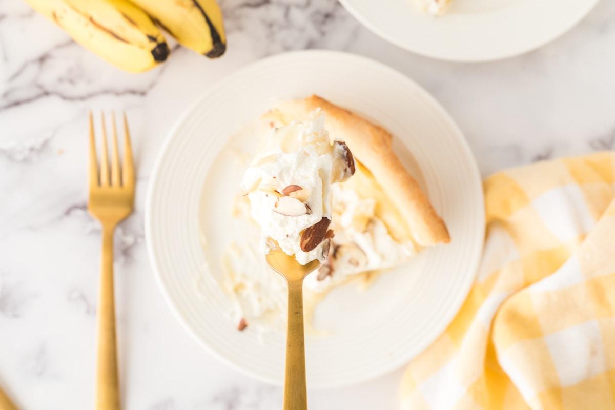 a bit of banana cream pie on a fork held over a slice of banana cream pie on a plate with almonds on top and bananas on a table