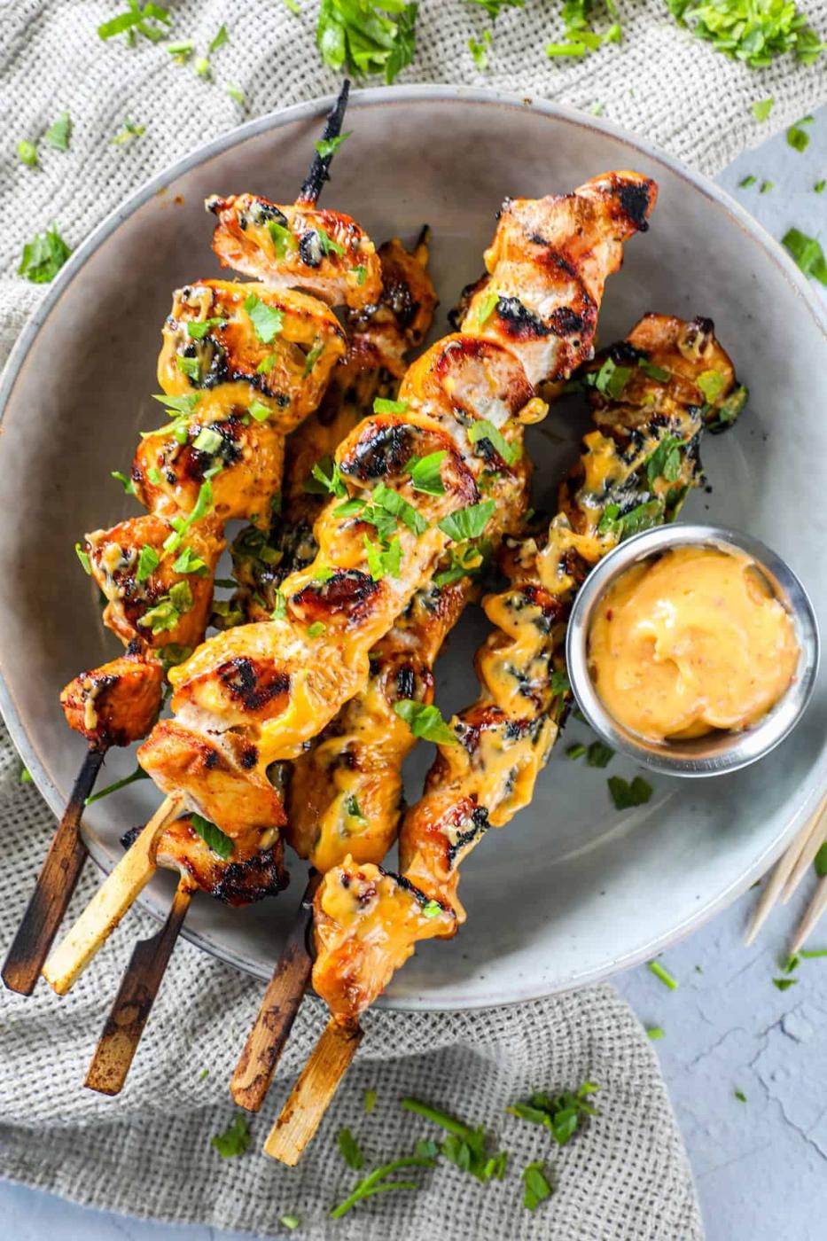 Grilled chicken skewers covered in a yellow sauce, garnished with chopped herbs, and served with a small bowl of dipping sauce on a white plate.