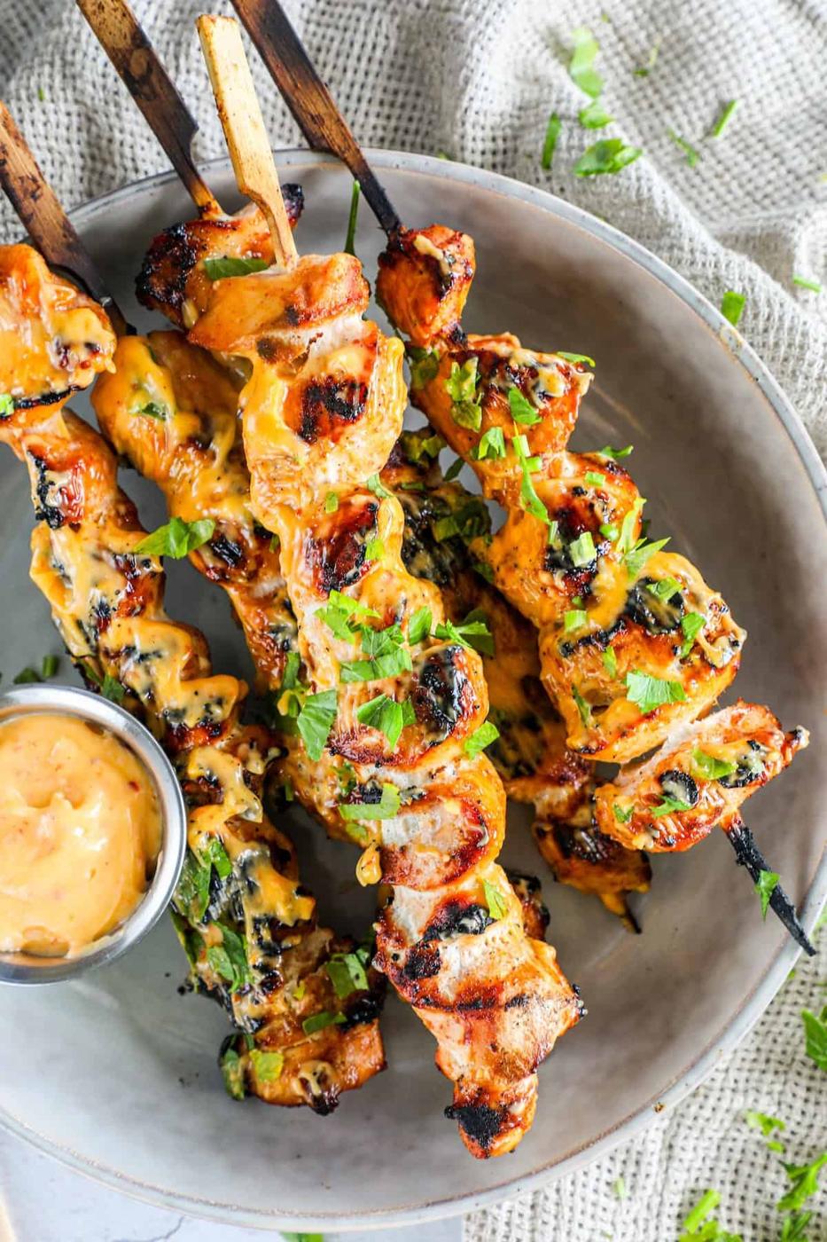 Grilled chicken skewers drizzled with sauce, garnished with chopped herbs, and served with a small dipping sauce in a bowl.