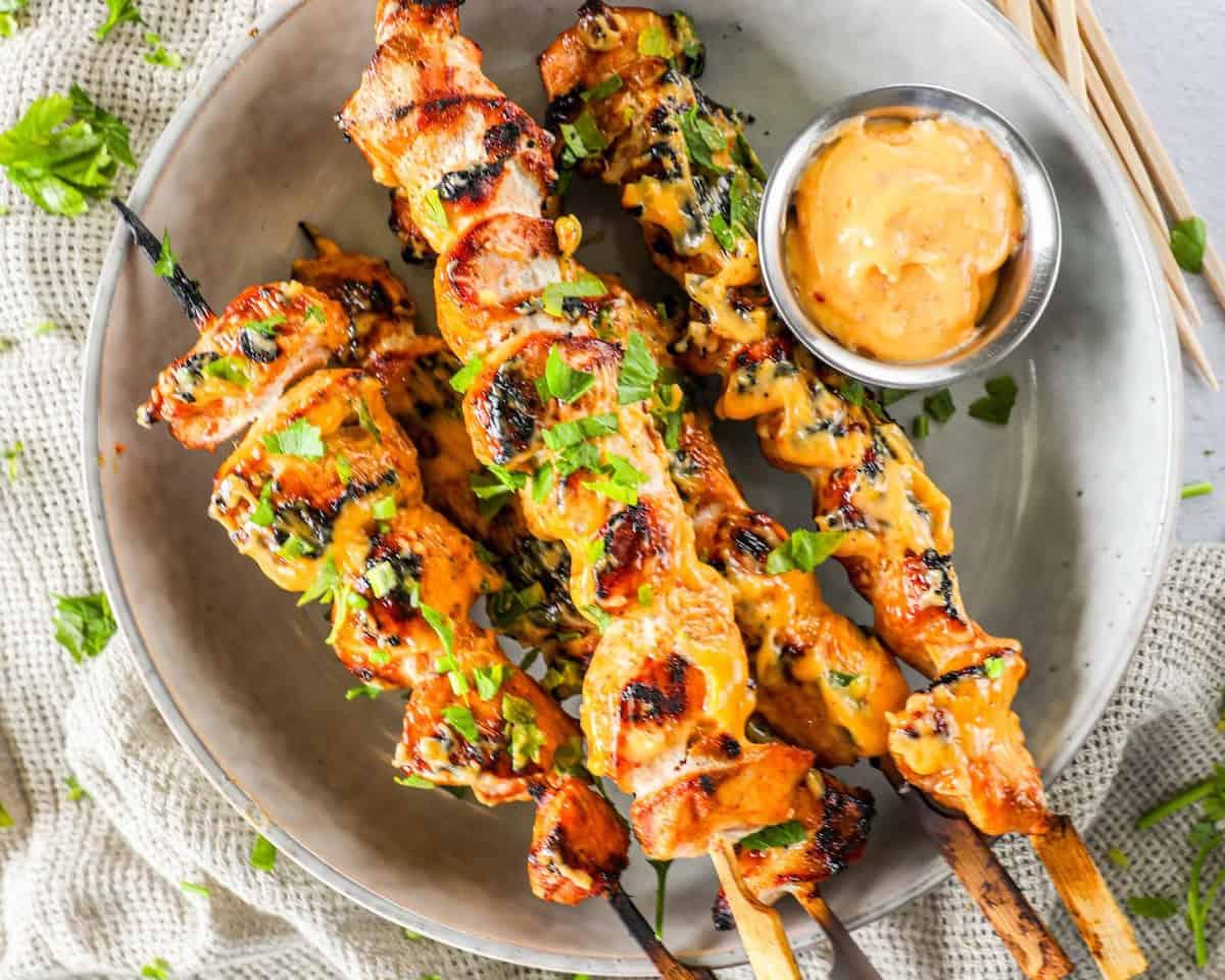 Plate of grilled chicken skewers garnished with parsley, accompanied by a small bowl of dipping sauce. The skewers are arranged on a beige plate placed on a textured fabric.