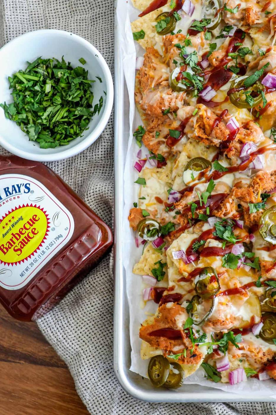 A tray of barbecue chicken nachos topped with jalapeños, red onions, and cilantro next to a bottle of barbecue sauce.