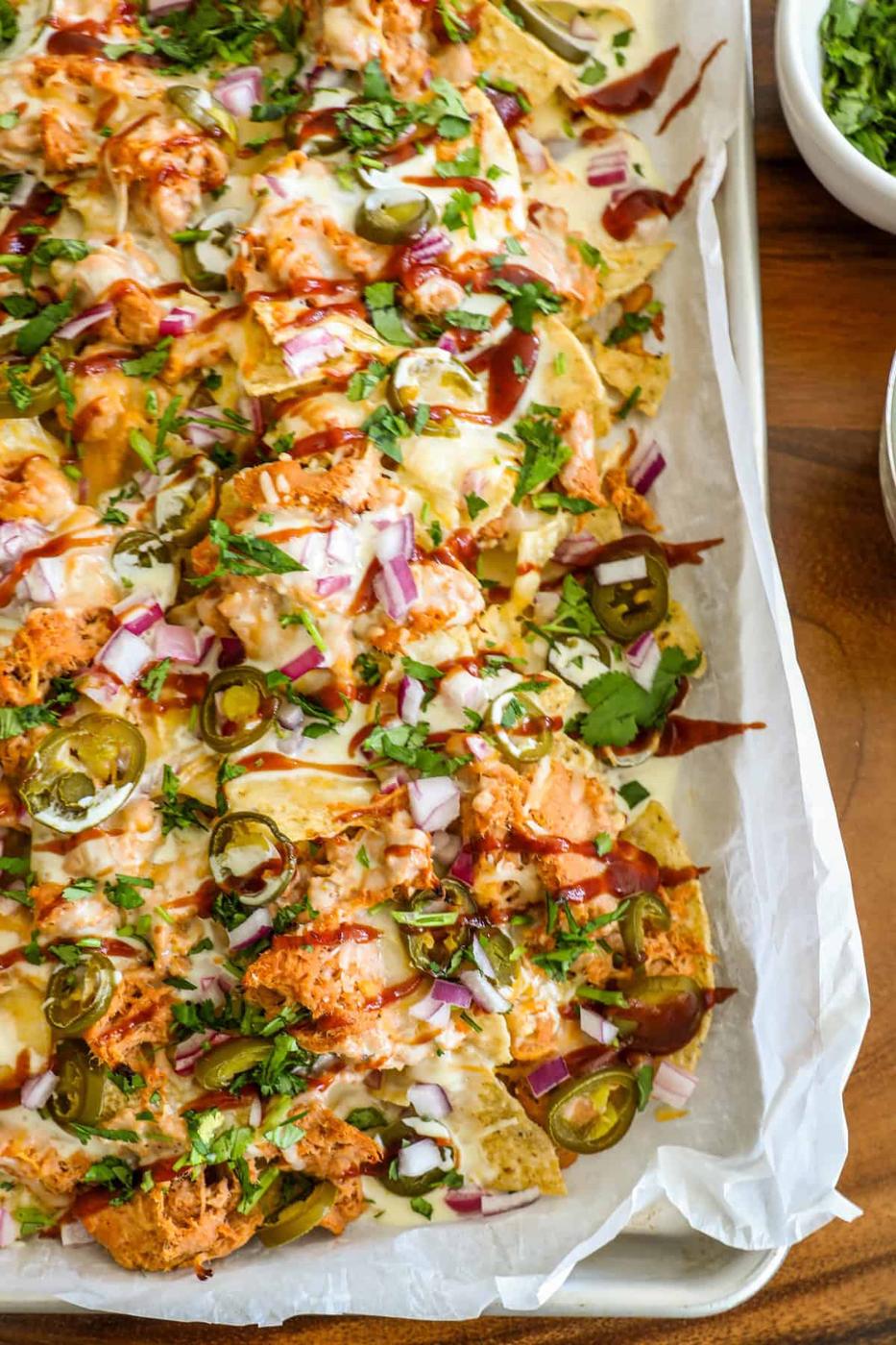 A tray of loaded nachos topped with melted cheese, shredded chicken, jalapeños, red onions, and a drizzle of sauces, served on a wooden table.