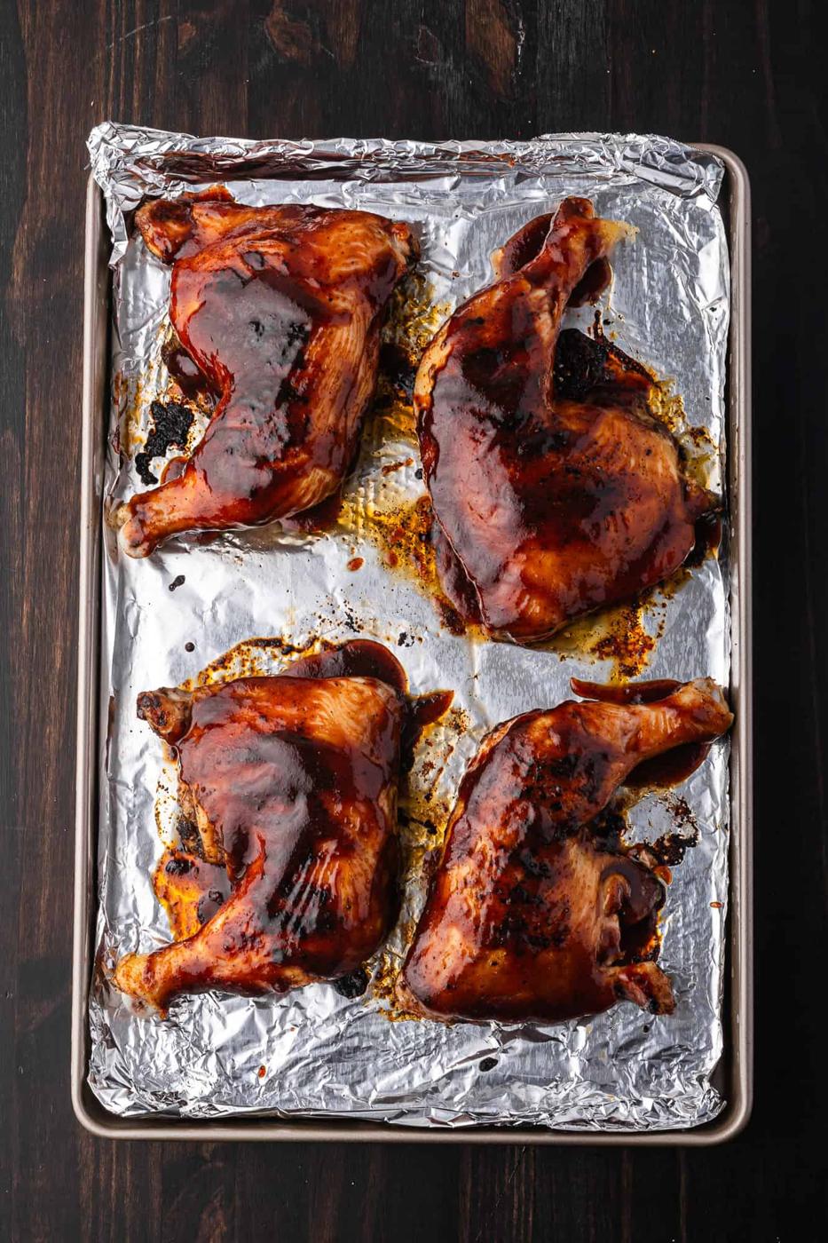 Four BBQ chicken legs with crispy skin and dark glaze rest on a foil-lined baking sheet, perfecting an easy BBQ recipe.