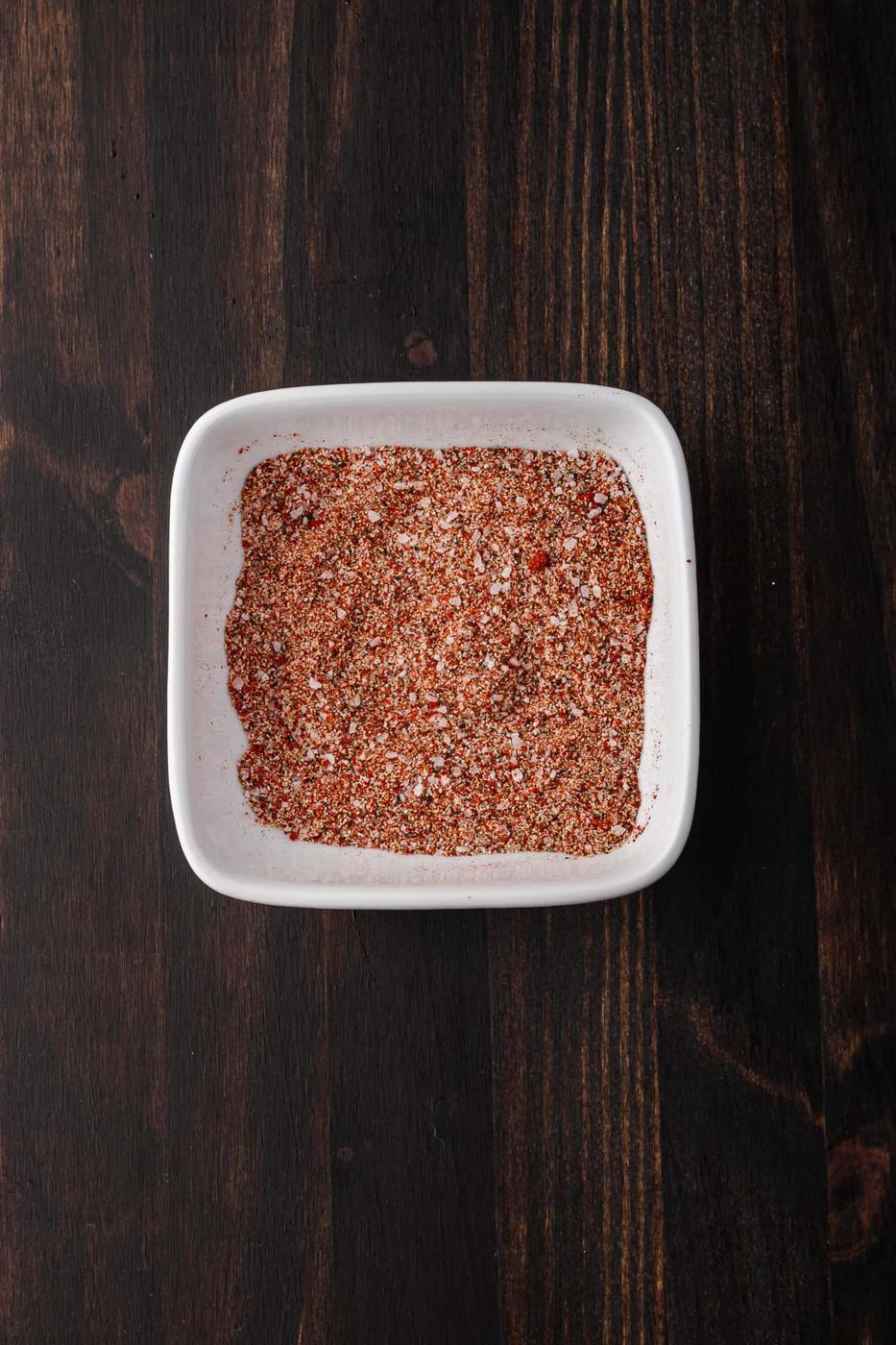 A white square dish filled with a blend of red and brown spices sits on a dark wooden surface, perfect for seasoning BBQ chicken.