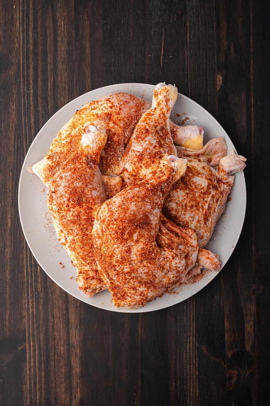 A plate of raw chicken quarters seasoned with spices, perfect for the BBQ, placed on a dark wooden surface.