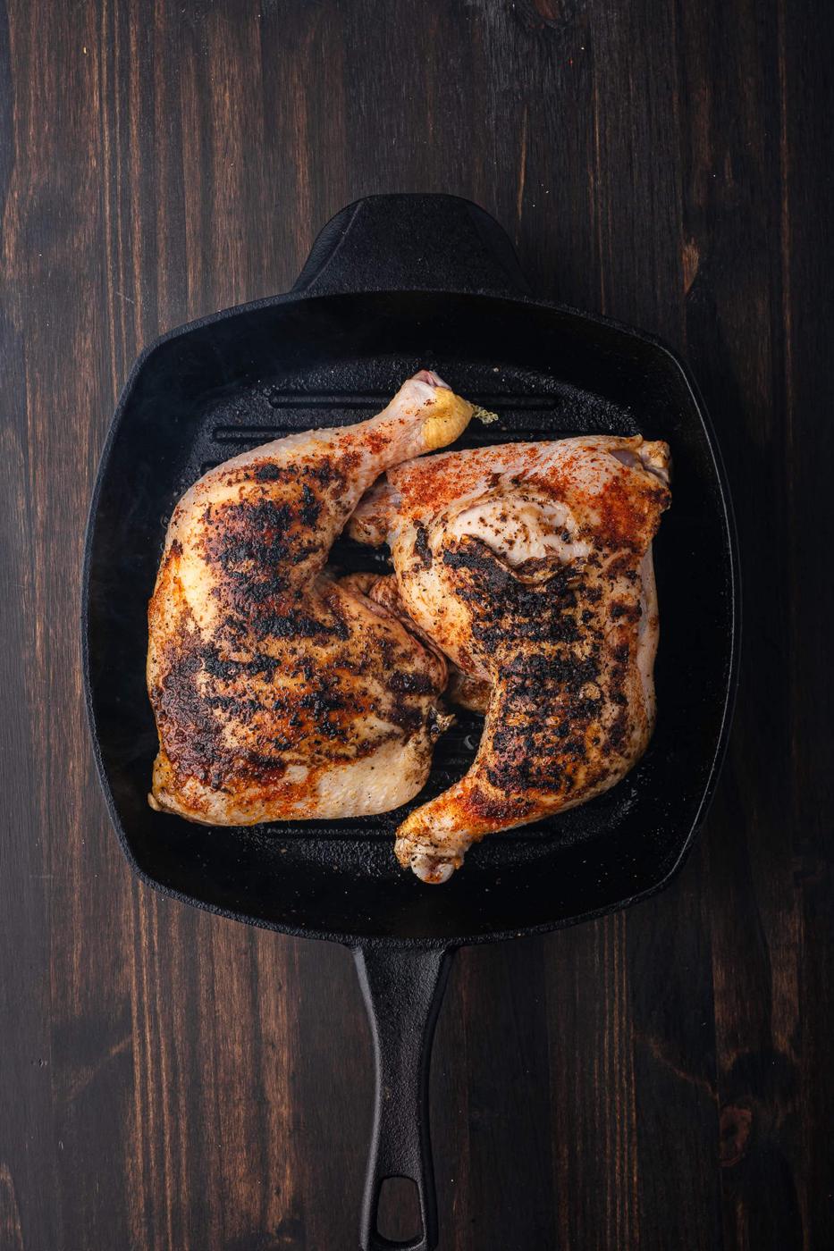 Two BBQ chicken quarters with crispy skin and seasoning rest in a black cast iron skillet on a dark wooden surface.