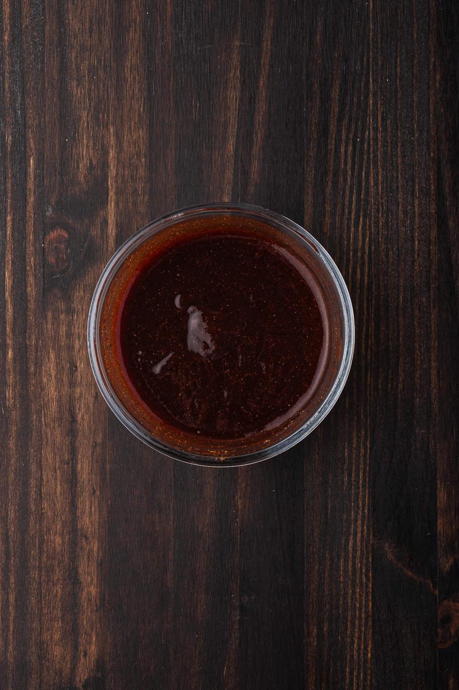 A small glass bowl filled with rich BBQ sauce rests on a dark wooden surface, perfect for slathering over grilled chicken quarters.
