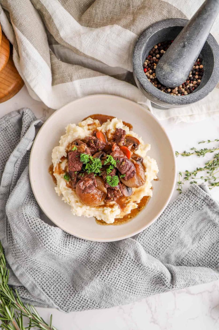 A plate of beef stew served over mashed potatoes, garnished with herbs, next to a mortar with peppercorns and sprigs of thyme.