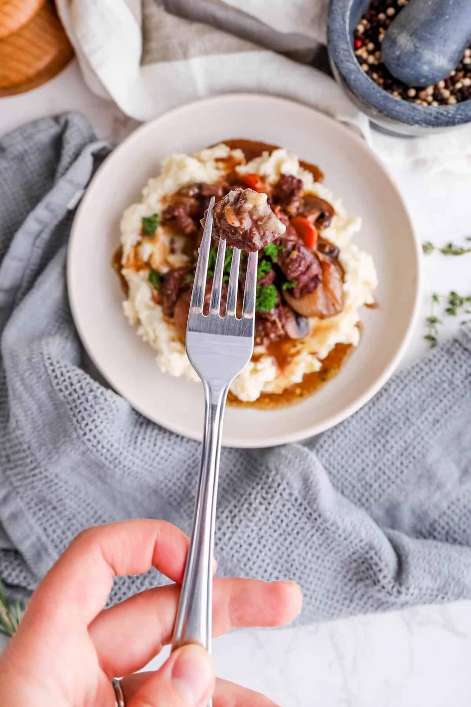 A person holding a fork with a piece of beef over a plate of beef stew and mashed potatoes, surrounded by a rustic kitchen setting.