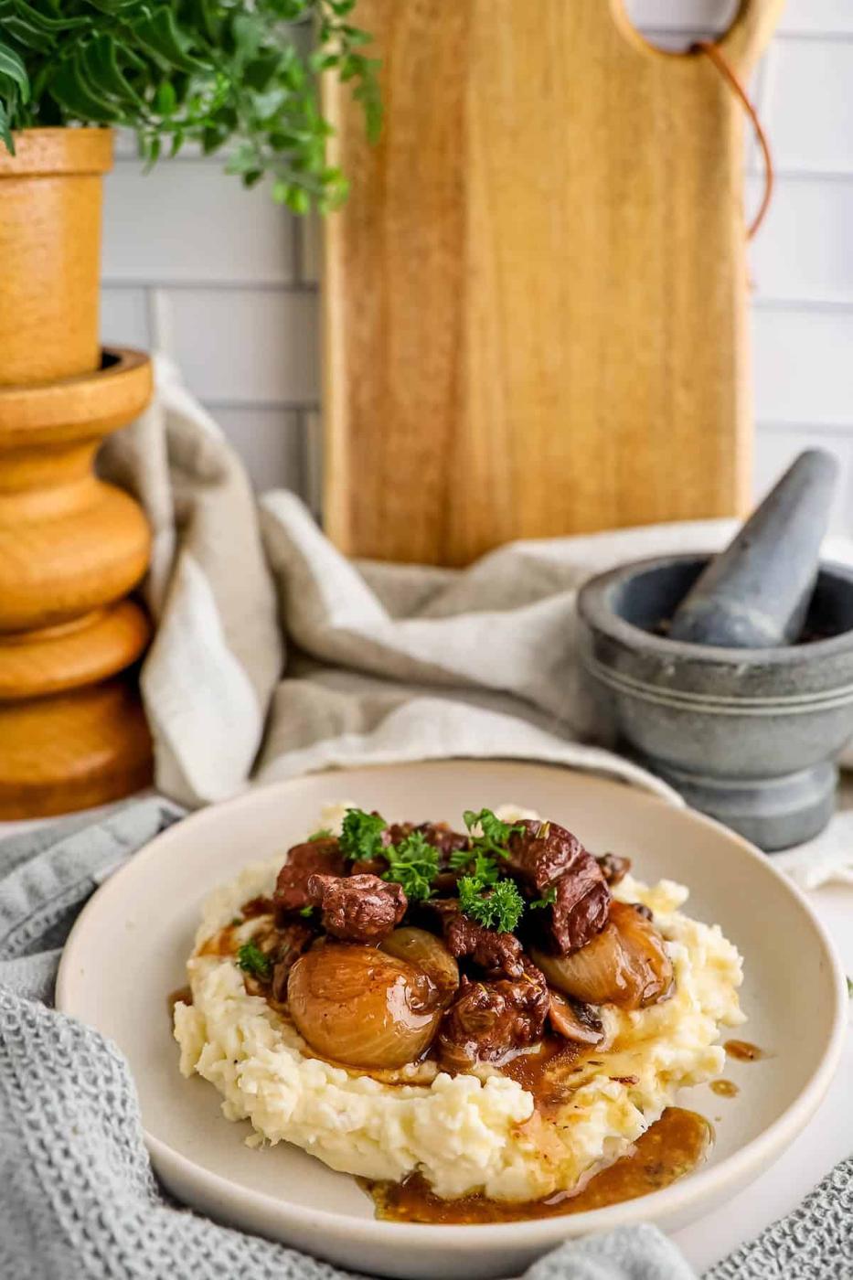 A plate of beef stew with onions over mashed potatoes, garnished with parsley, beside a wooden pepper mill and mortar on a kitchen counter.
