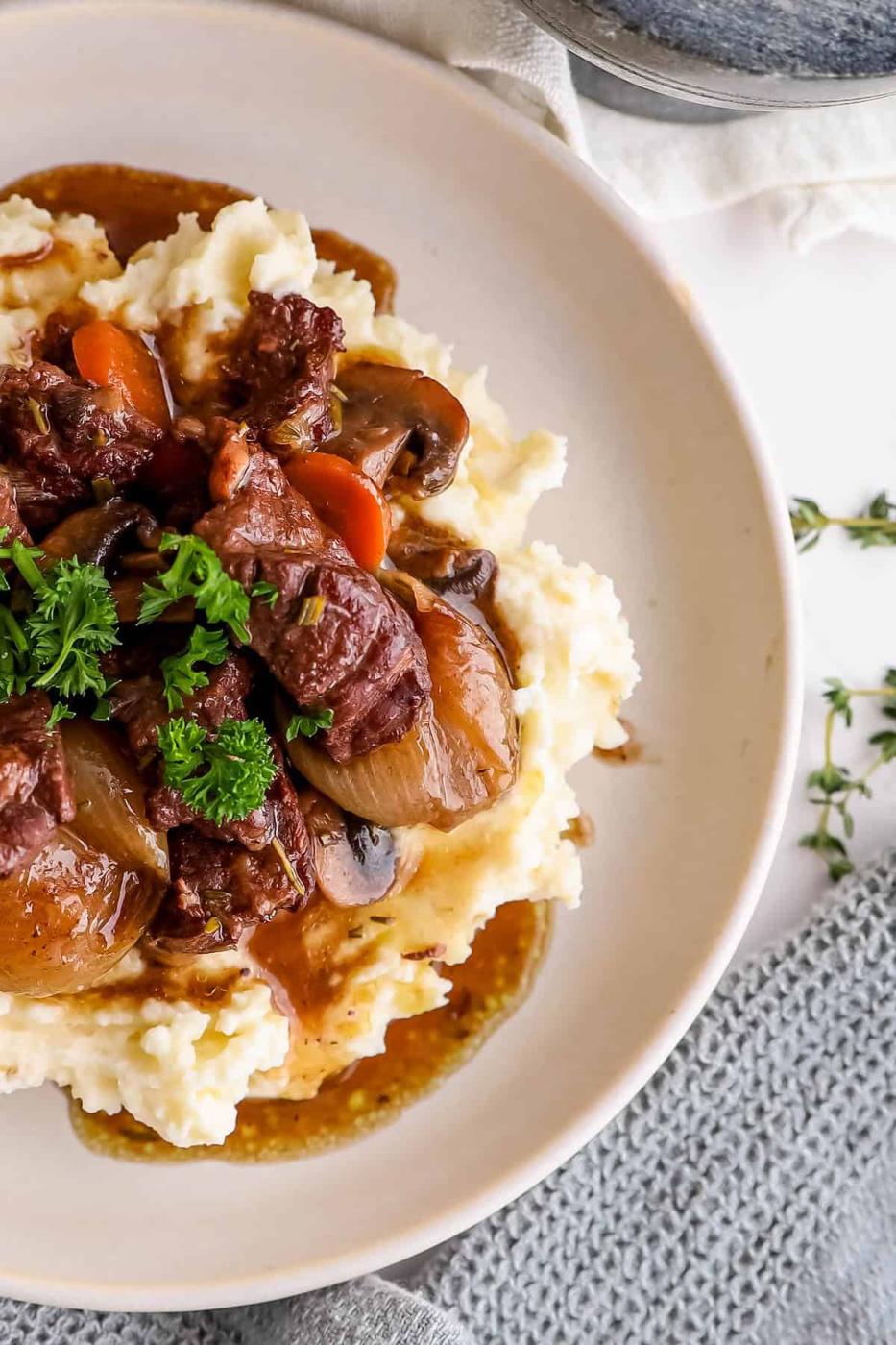 A plate of beef stew served over mashed potatoes, garnished with parsley, accompanied by thyme on a light background.