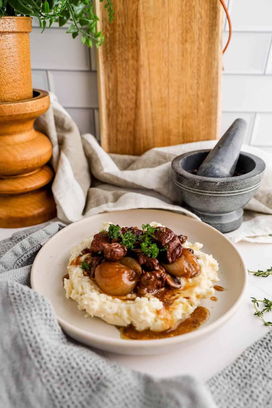 A plate of beef stew with whole mushrooms served over mashed potatoes, garnished with parsley, set on a wooden kitchen counter.