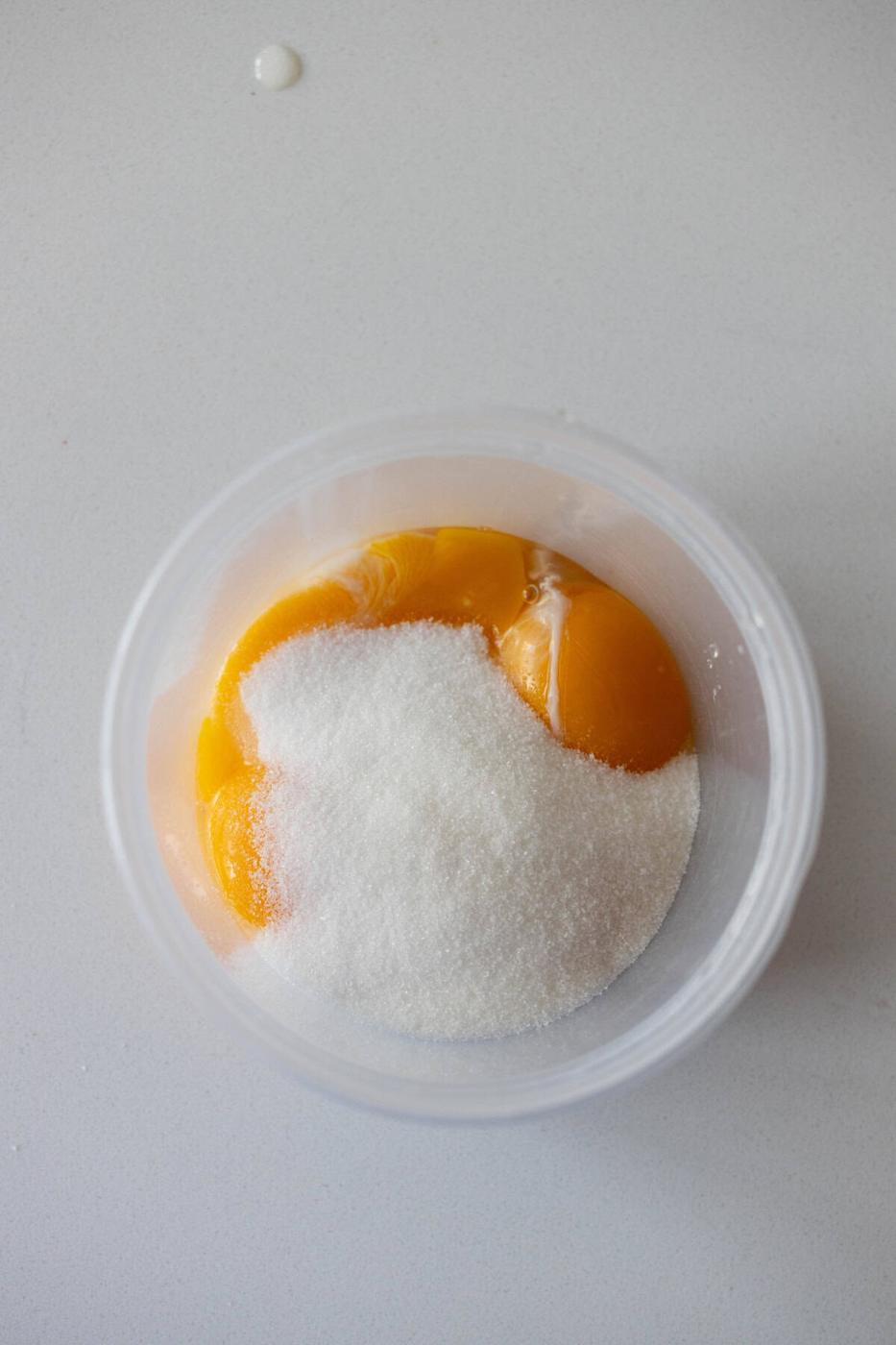 A clear container holding several egg yolks and a pile of granulated white sugar on a light-colored surface, ready to be transformed into a luscious Berries and Creme Anglaise.
