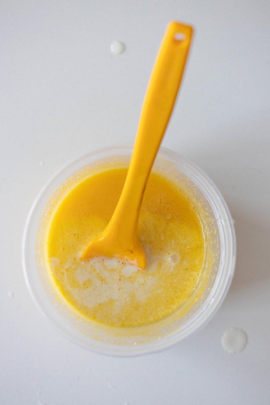 A yellow spoon in a clear plastic container with beaten eggs and a touch of Berries and Creme Anglaise on a white surface, seen from above.