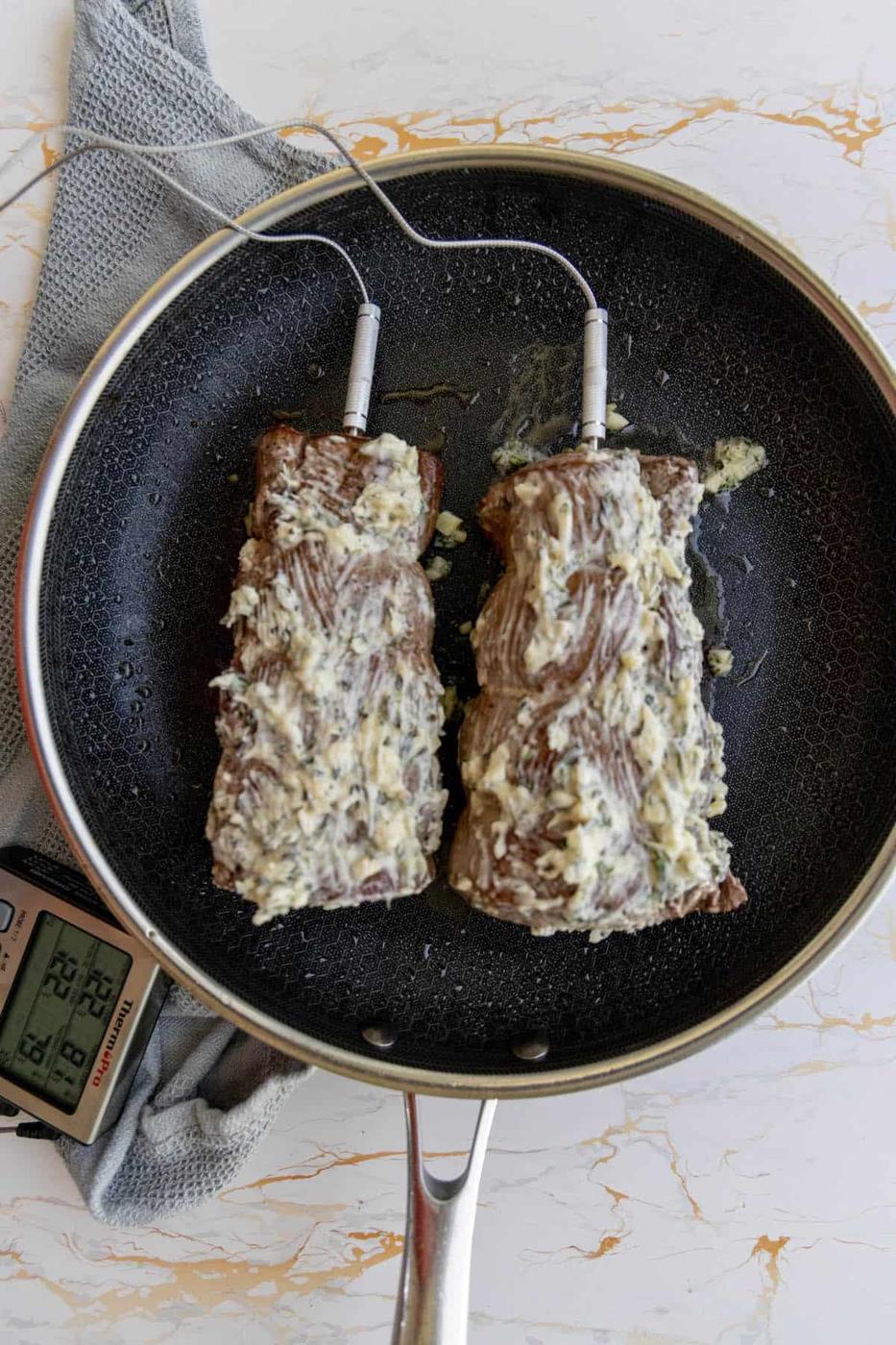 Two garlic herb beef tenderloins sizzling in a frying pan with melted cheese on top.