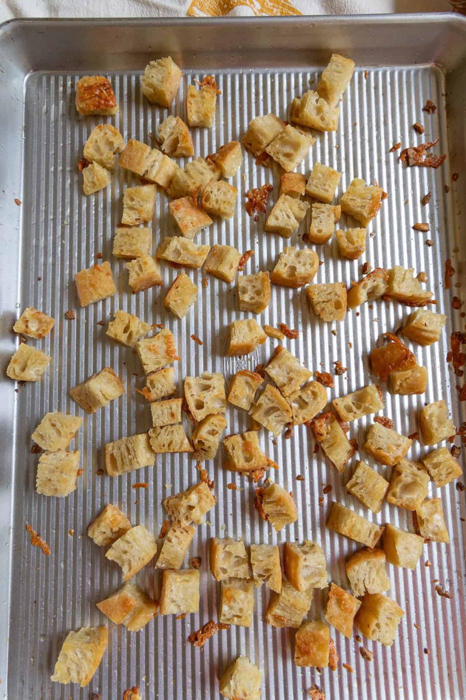 Cubed pieces of toasted bread are scattered on a ridged metal baking sheet, some with melted cheese and a drizzle of Caesar dressing on top.