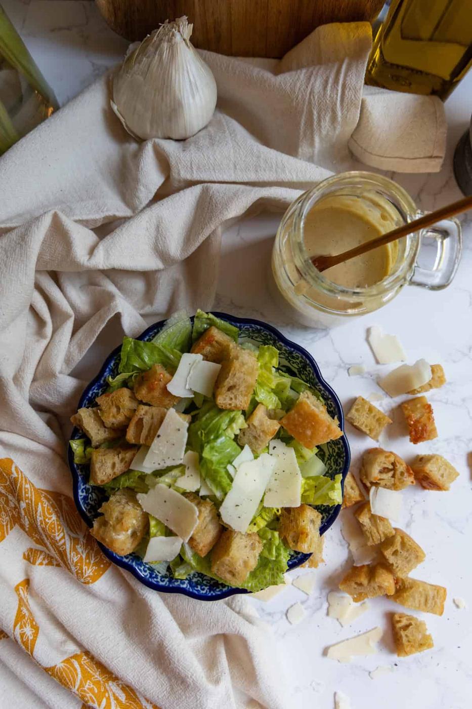 A bowl of Caesar salad with croutons and shaved parmesan sits on a cloth, next to a garlic bulb, a jar of Caesar dressing with a spoon, and scattered croutons and cheese.