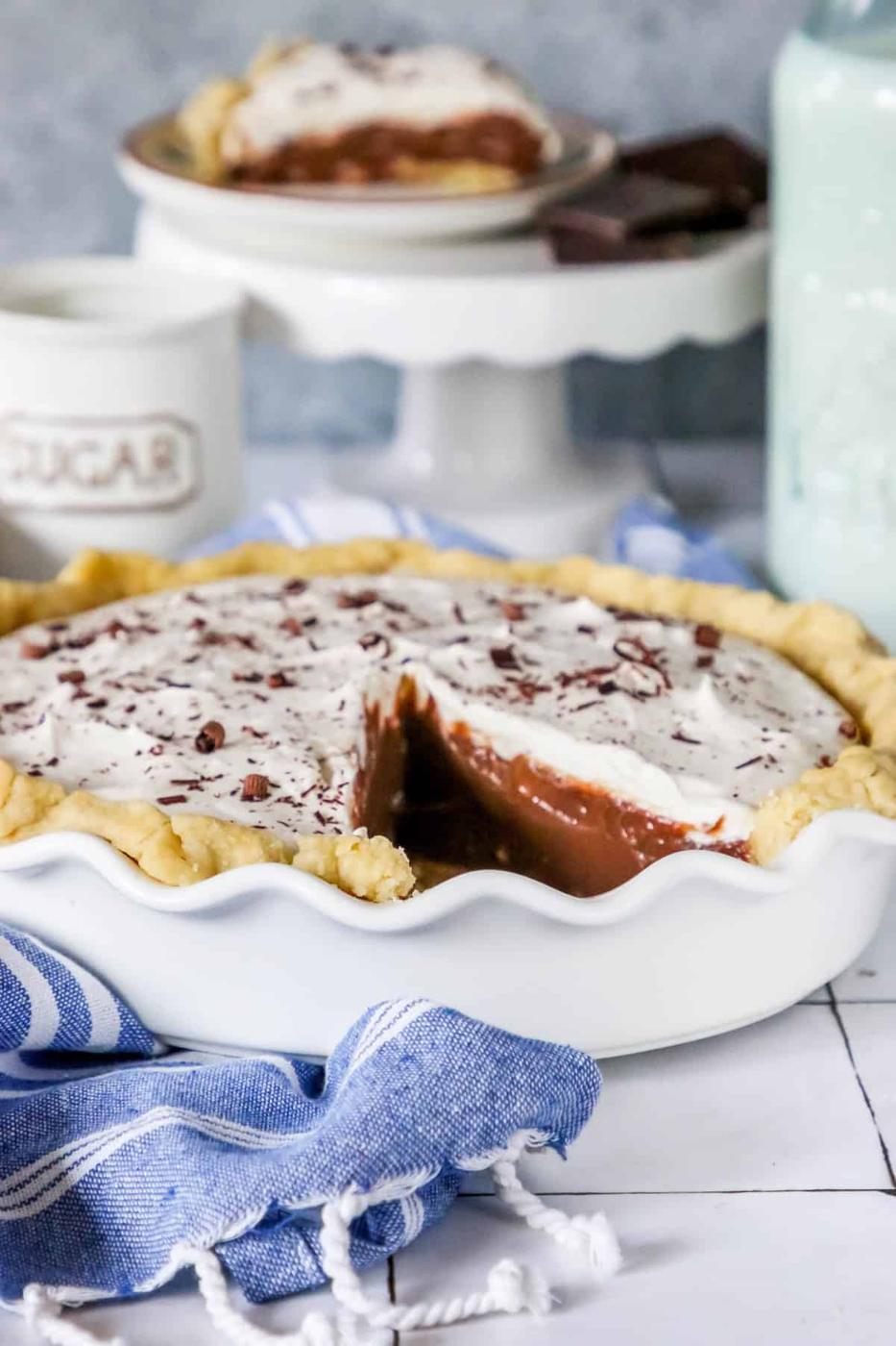 A whole chocolate pie with one slice removed, revealing its smooth filling, garnished with whipped cream and chocolate shavings, set on a white dish beside a sugar container.