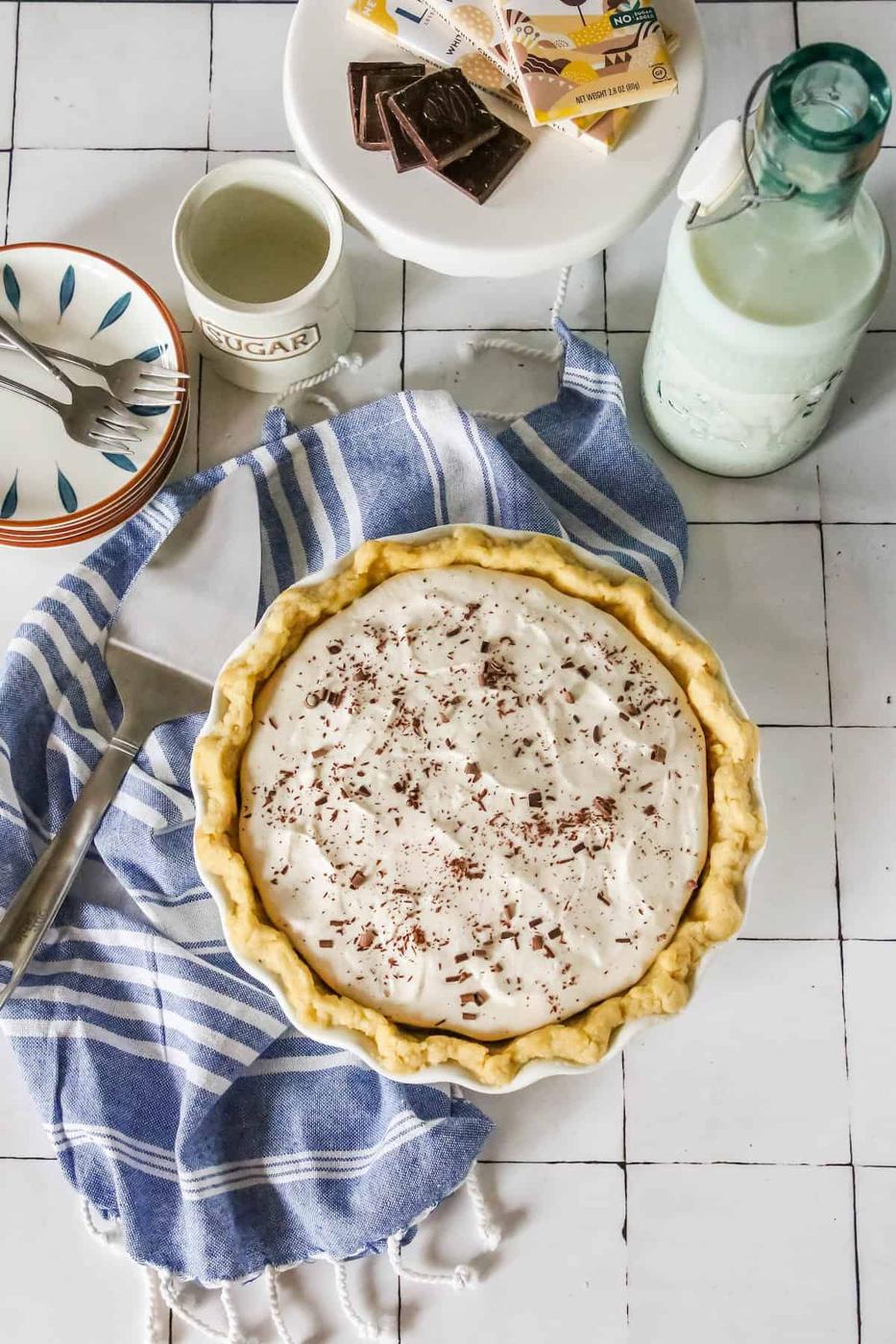 A freshly made chocolate pie on a blue-striped towel with a milk bottle, a sugar bowl, chocolate pieces, and a floral plate nearby on a tiled surface.