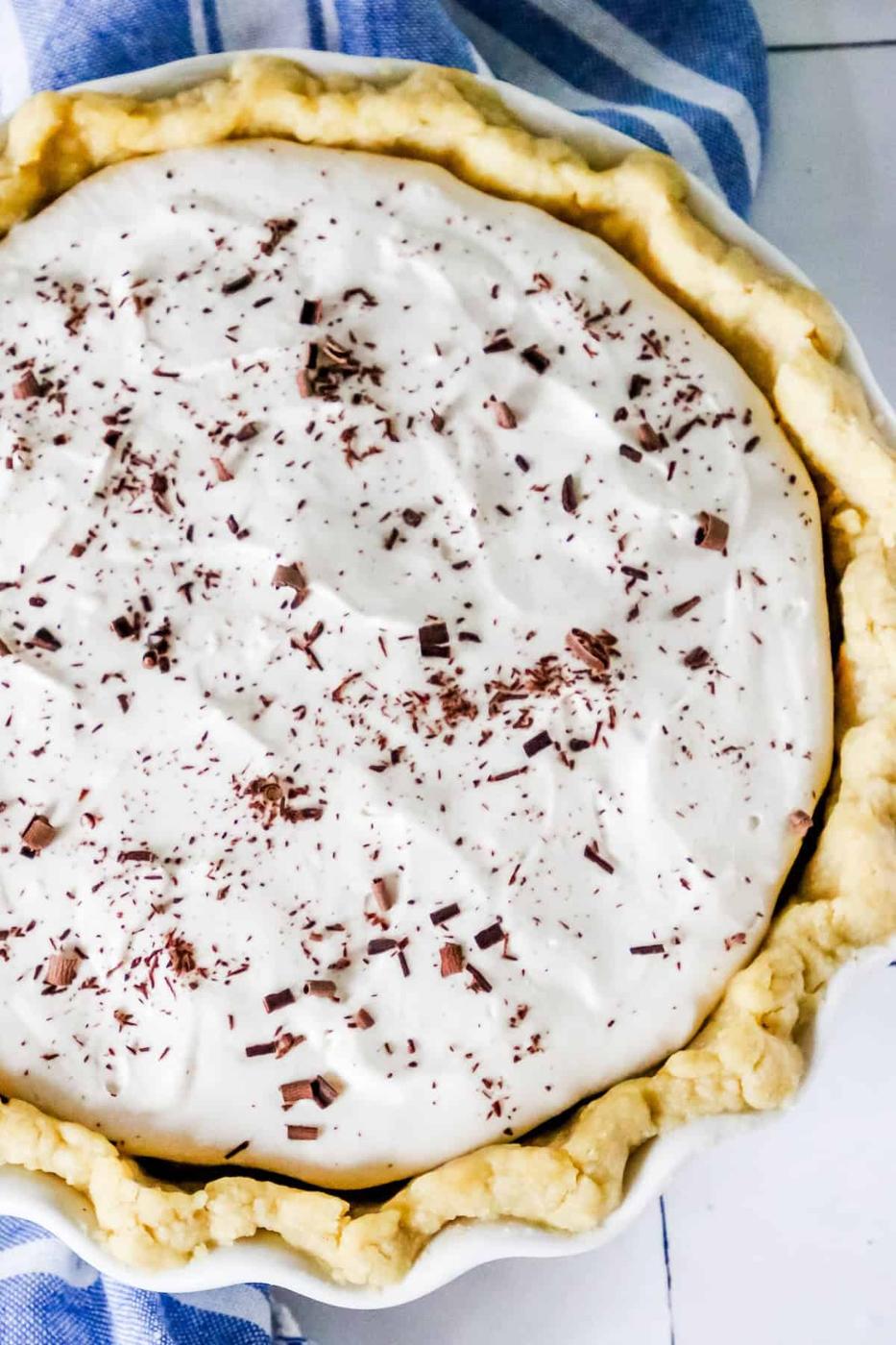 A homemade pie topped with white frosting and sprinkled with chocolate shavings, presented on a blue-striped cloth.