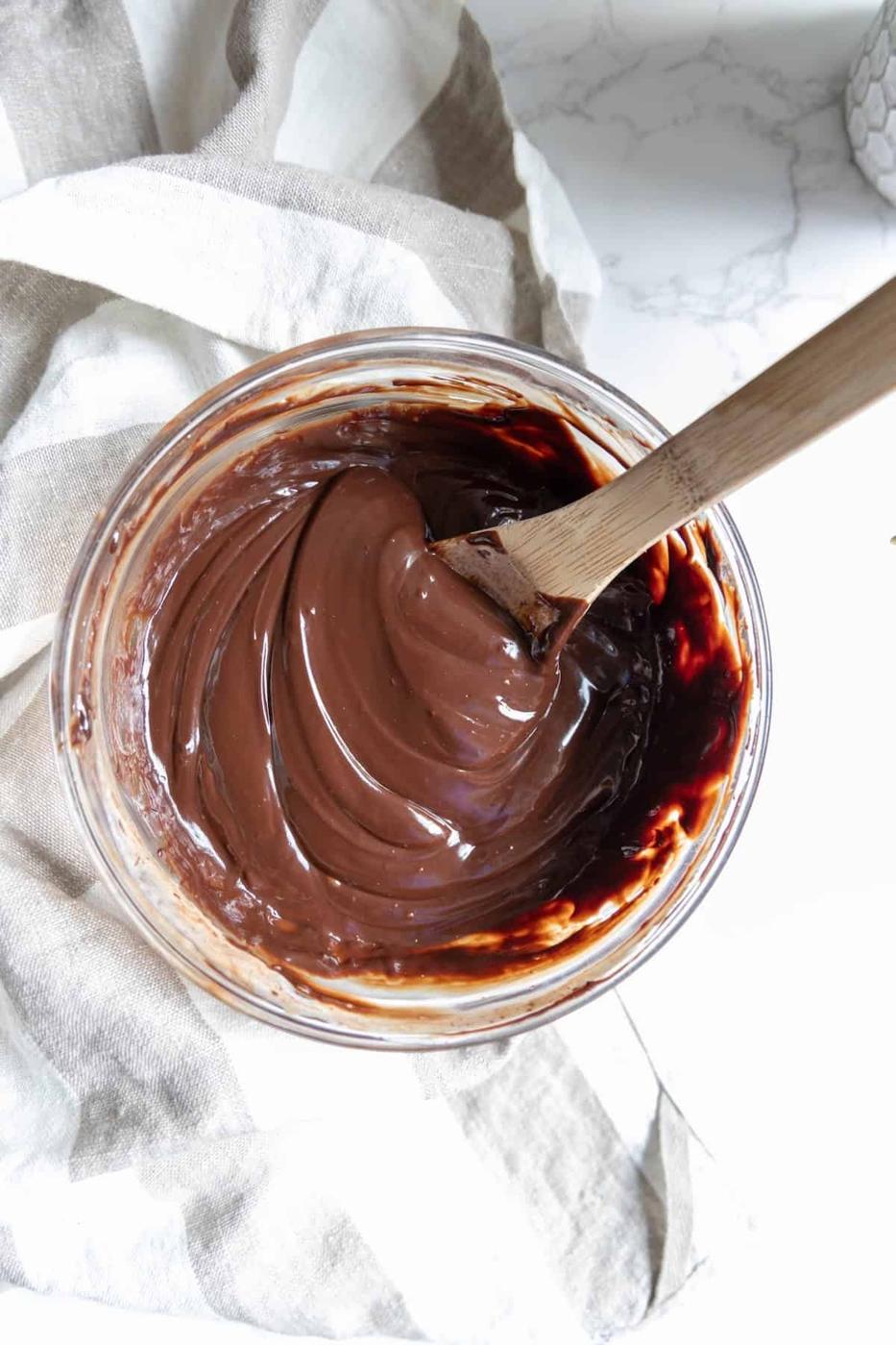 A glass bowl of smooth, glossy chocolate mixture for chocolate truffles sits with a wooden spoon resting inside, placed on a striped cloth.