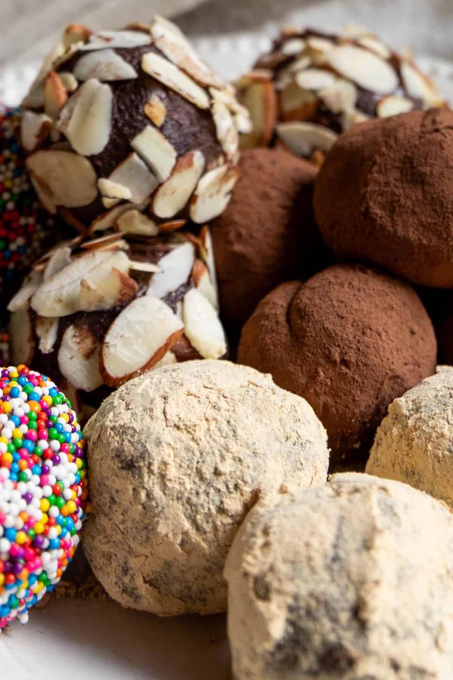 Assorted chocolate truffles are covered in sliced almonds, cocoa powder, powdered sugar, and colorful sprinkles, all beautifully arranged on a plate.