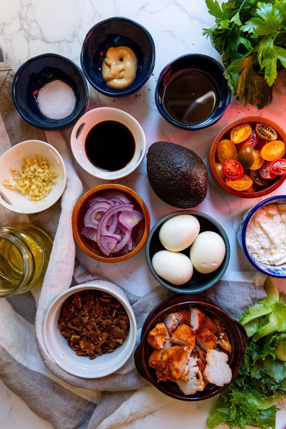 A flat lay of the best Cobb salad ingredients, featuring eggs, avocado, grilled chicken, bacon, red onion, grape tomatoes, cheese, sauces, oil, salt, minced garlic, and fresh greens on a marble surface.