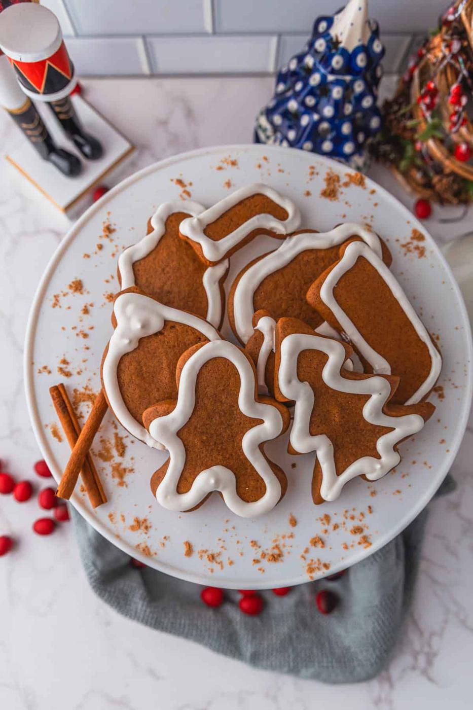 A plate of gingerbread cookies with white icing, shaped like a mitten, stocking, gingerbread person, bell, and tree sits invitingly on a table adorned with cinnamon sticks and red berries.