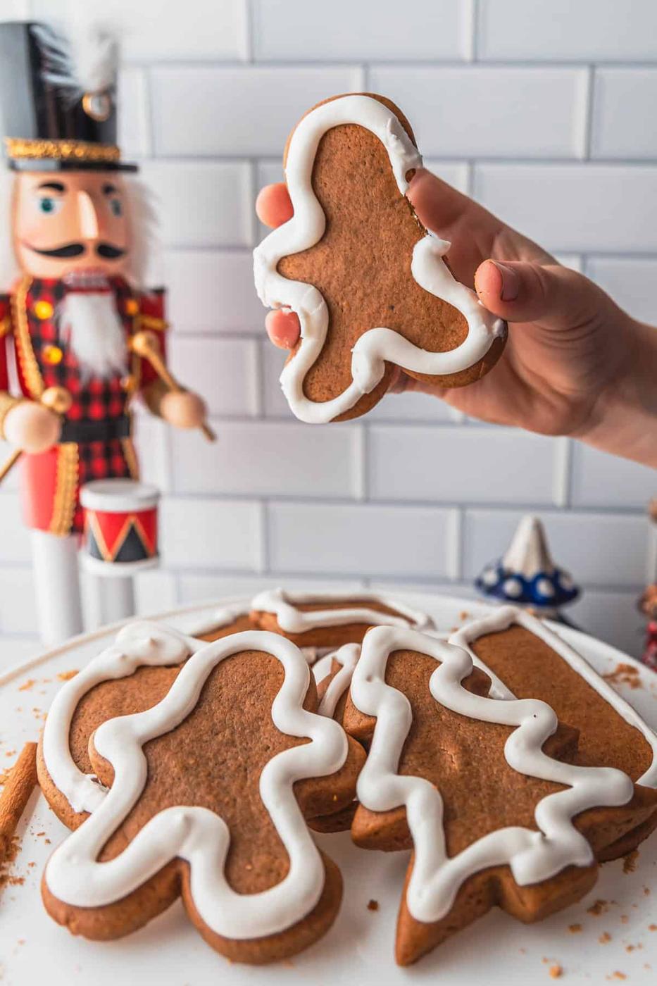 A hand holds a gingerbread cookie with white icing, hovering above a plate of gingerbread cookies, while a nutcracker figure stands watchful in the background.