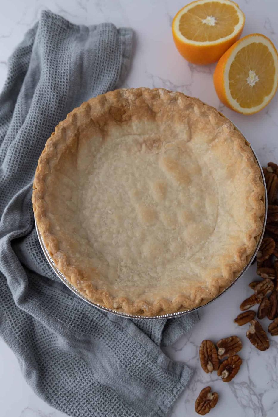 A baked pie crust in a tin next to a gray cloth, halved orange, and scattered pecans on a marble surface.