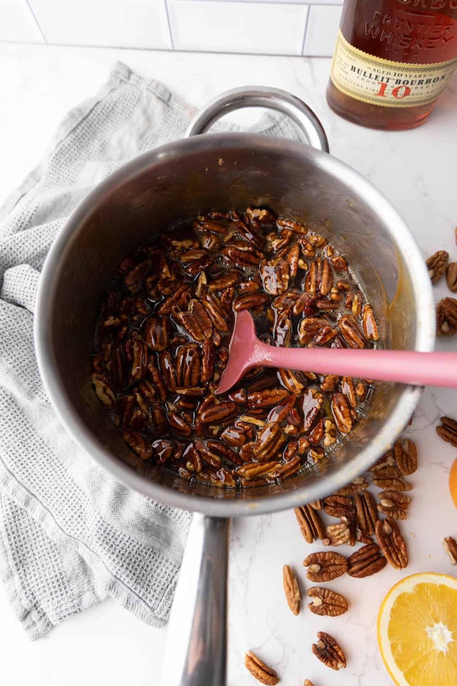 A saucepan of pecans coated in a brown liquid on a marble surface, with a pink spatula inside. A bottle and orange slices are visible in the background.
