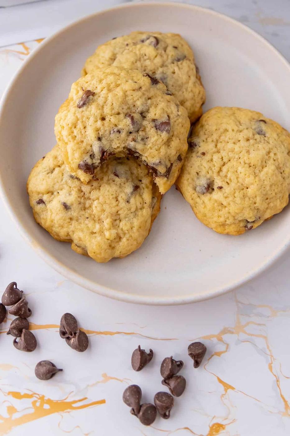 A white plate holds four sourdough chocolate chip cookies; one has a bite taken out. Scattered chocolate chips lie on the marble-like surface around the plate.