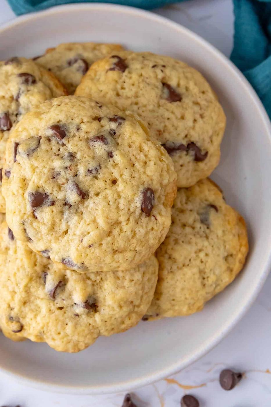 A close-up of a plate with a stack of sourdough chocolate chip cookies on a white surface. A few chocolate chips are scattered beside the plate. A teal cloth is partially visible in the background.