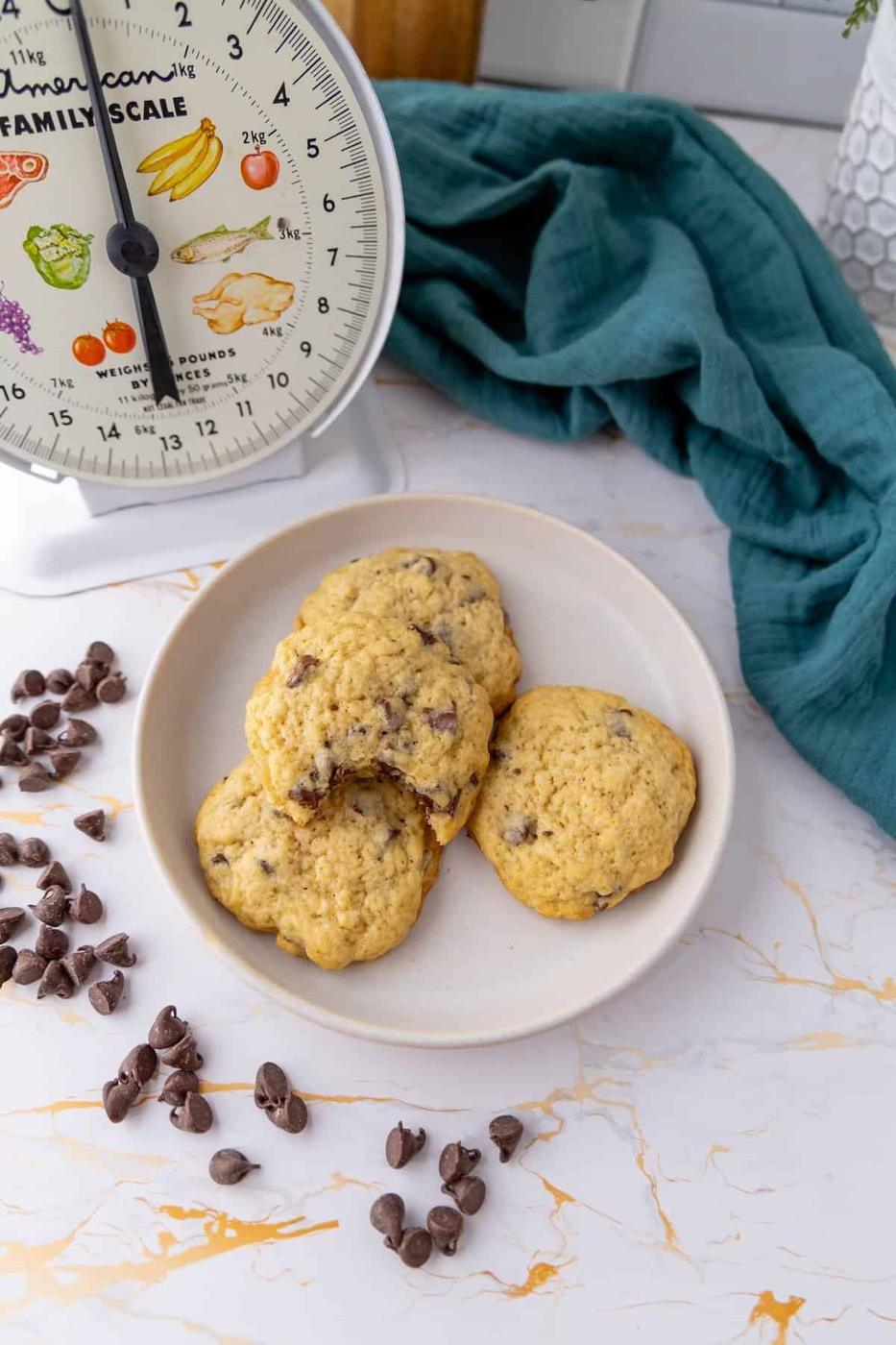 A plate of sourdough chocolate chip cookies is placed beside scattered chocolate chips and an old-fashioned scale on a marble countertop. A teal cloth is in the background.