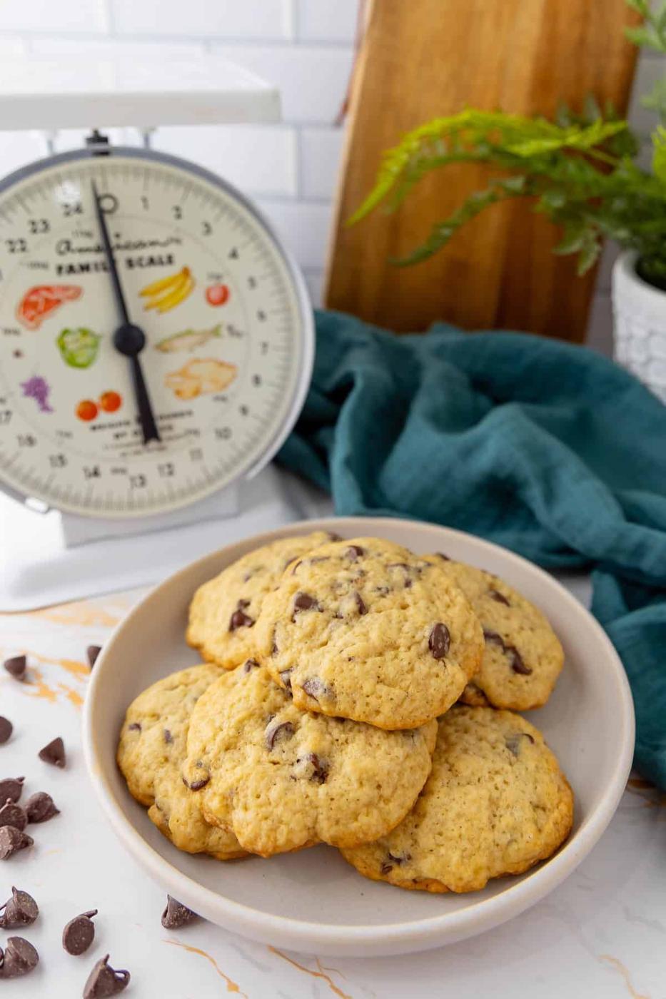 A plate of sourdough chocolate chip cookies on a marble countertop, with a vintage kitchen scale and a green cloth in the background. A plant in a white pot is also visible.
