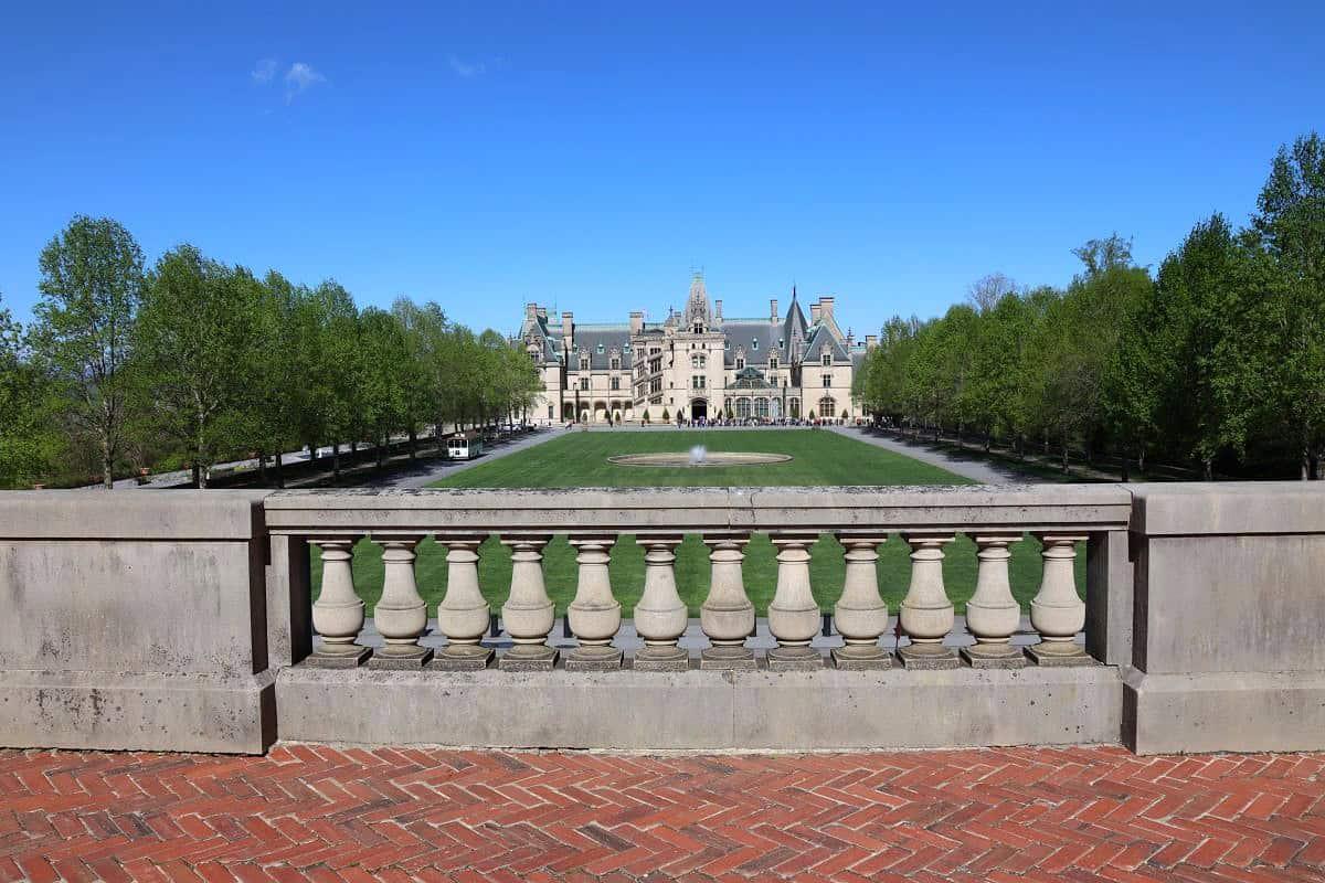 A large historic mansion with formal gardens is viewed from behind a stone balustrade, with a fountain and rows of trees lining the lawn under a clear blue sky.